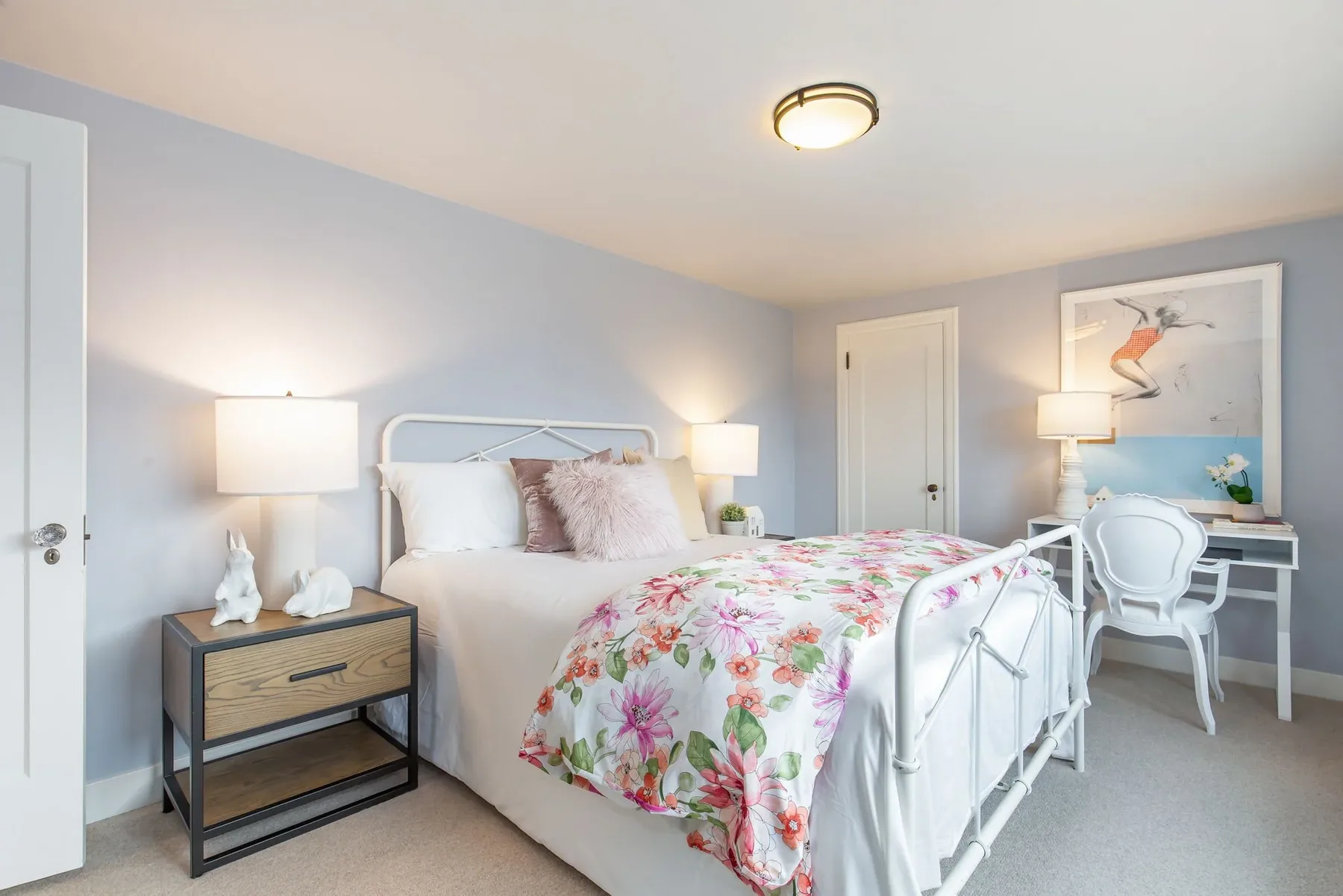 Guest bedroom staged with lavender walls, a white iron bed, floral bedding, and a writing desk beside the window