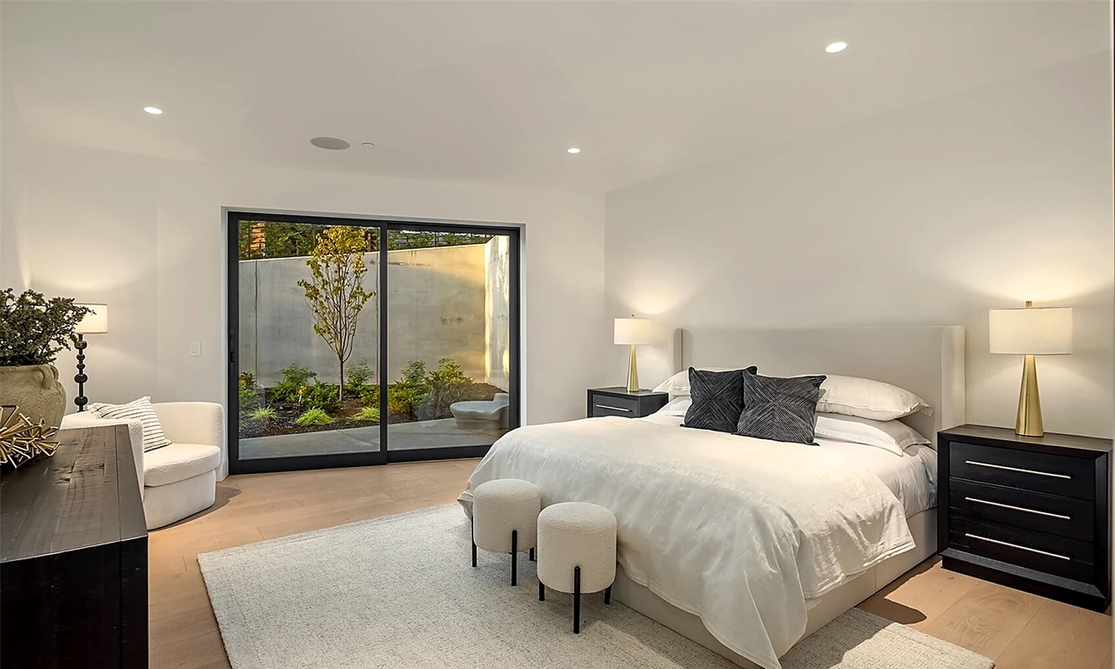 Serene staged bedroom with an upholstered bed, brass lamps, dark nightstands, and a sliding glass door to a landscaped courtyard