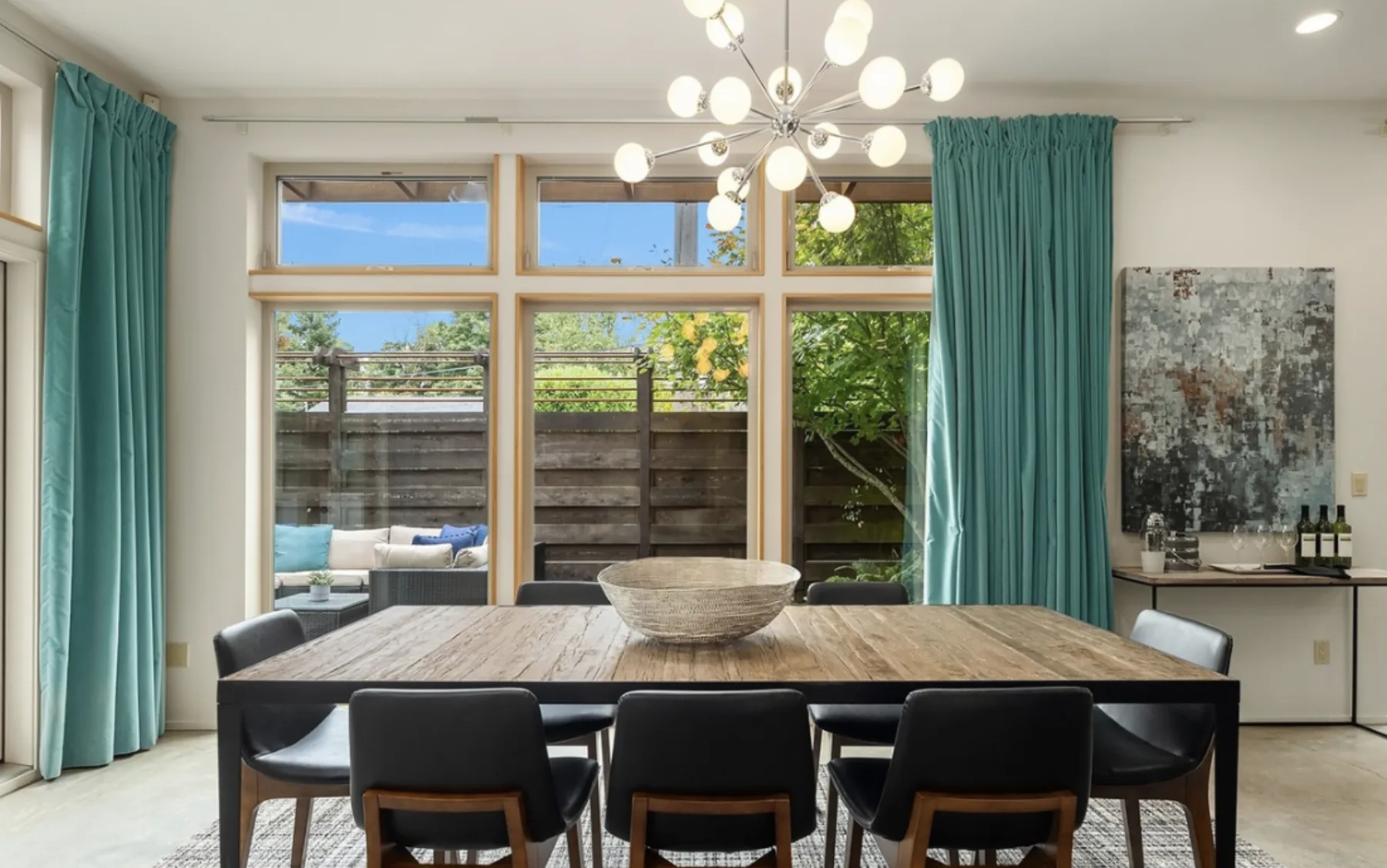 Dining room staged with a wood table, black leather chairs, a sputnik chandelier, and teal curtains beside windows to the courtyard