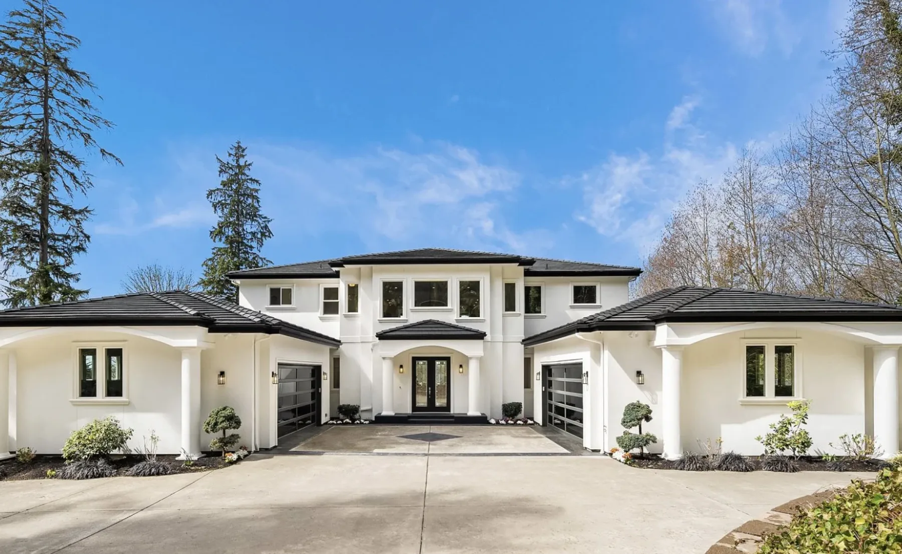 Seattle luxury home staging featuring a white contemporary estate with a tile roof, columned entry, and symmetrical garages among the trees