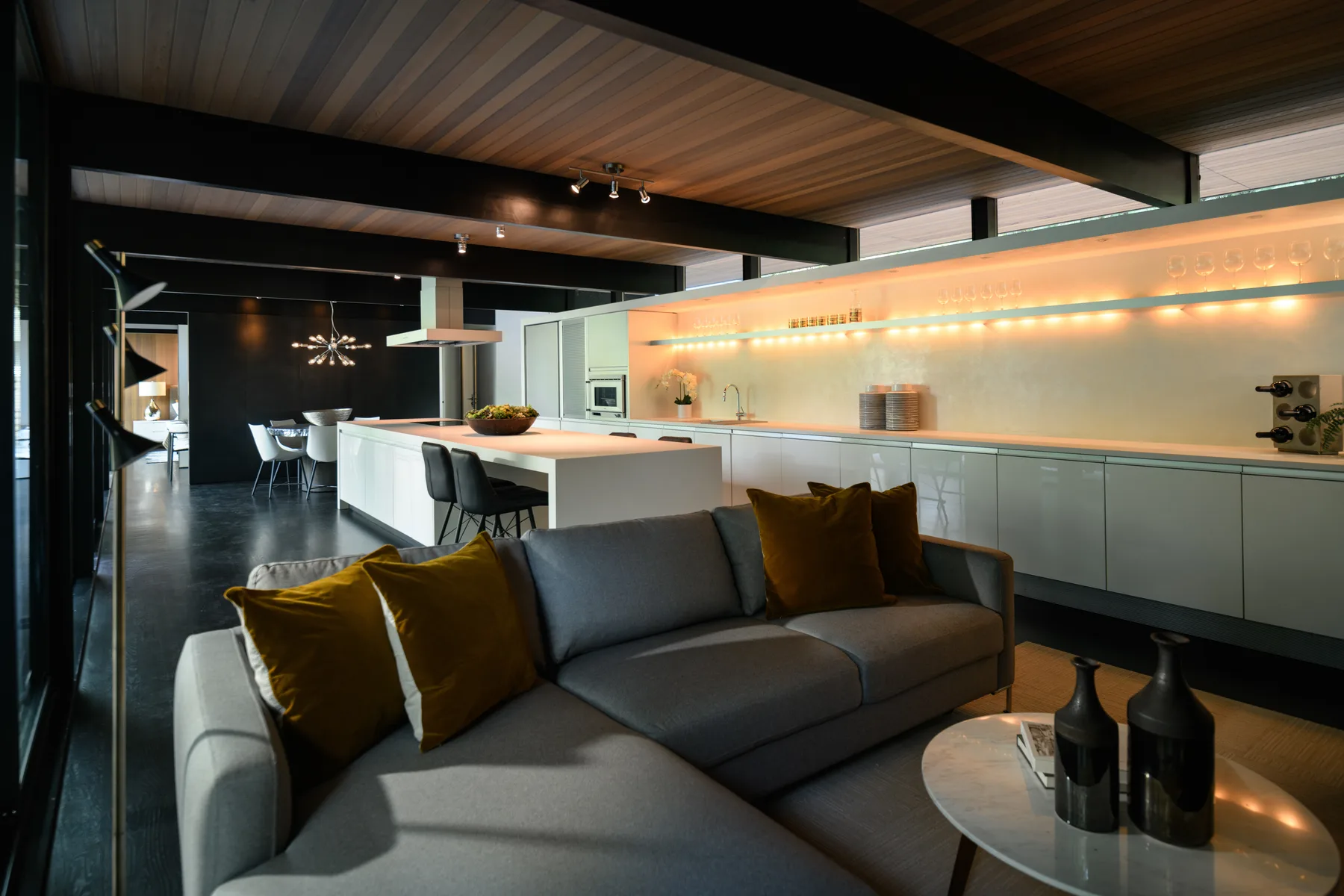 Open-plan great room with a grey sectional, walnut ceiling, and a backlit kitchen beyond