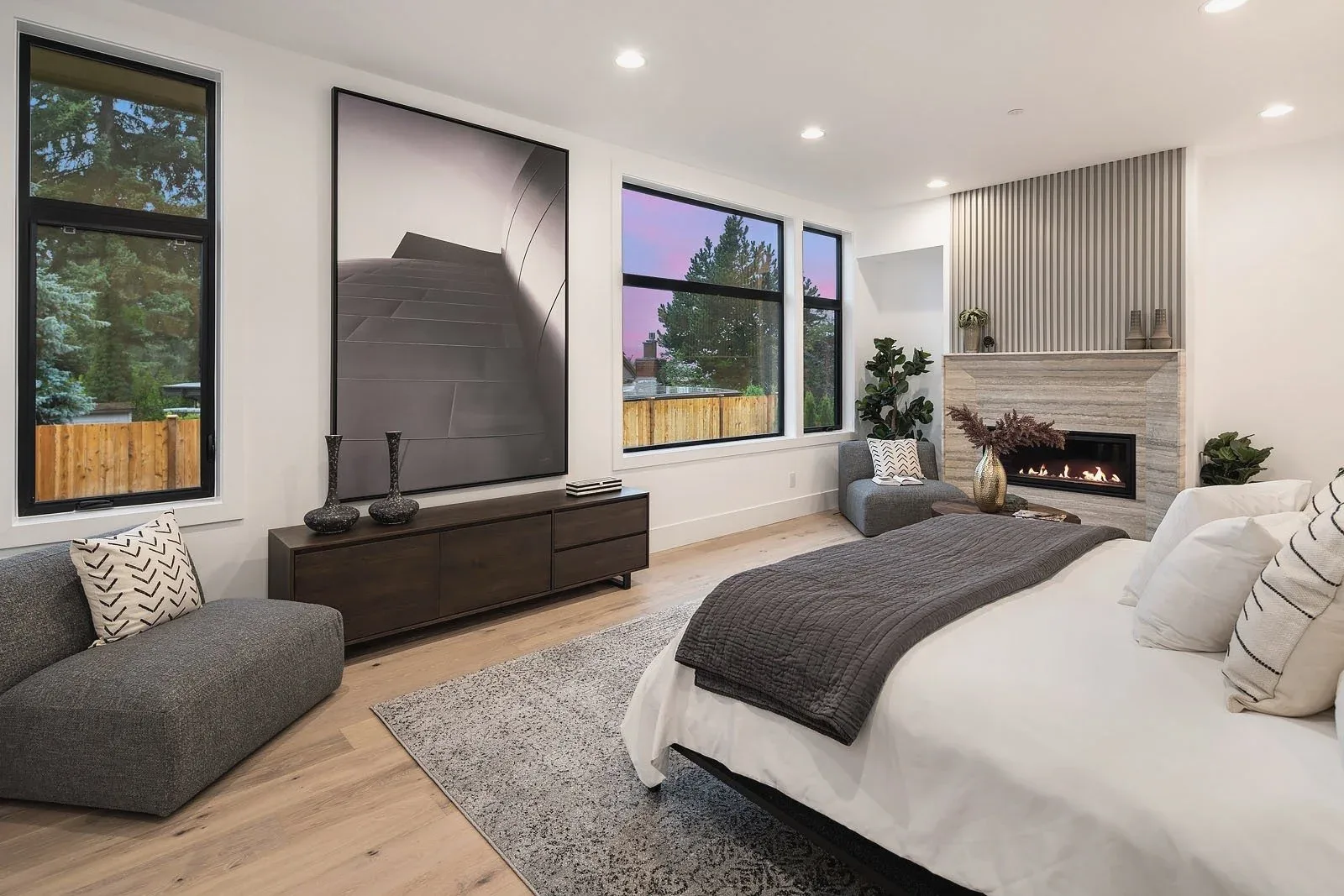Modern primary bedroom staged with a linear fireplace, layered textiles, and a cozy seating nook by the windows