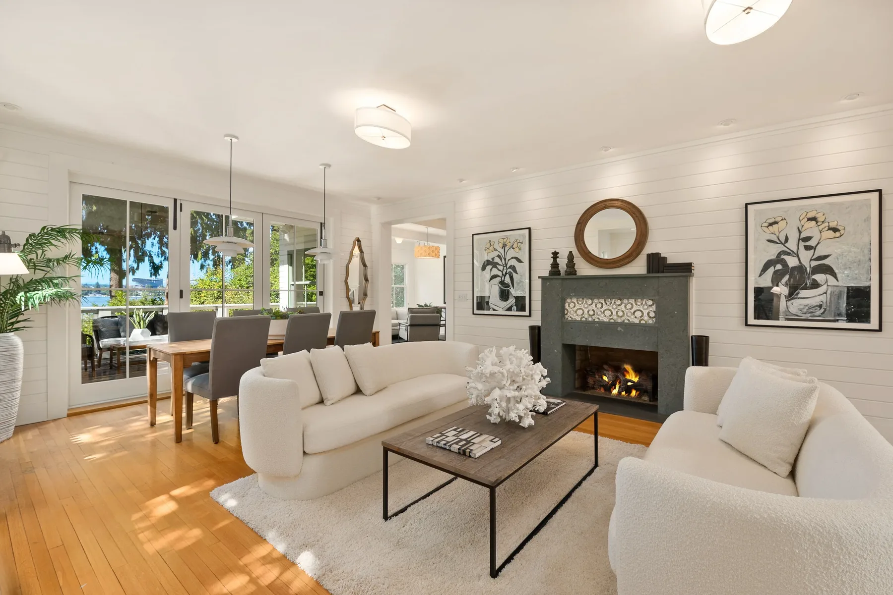Living room staged with a curved white sofa, green tile fireplace, round wood mirror, and botanical art opening to a lake-view dining area