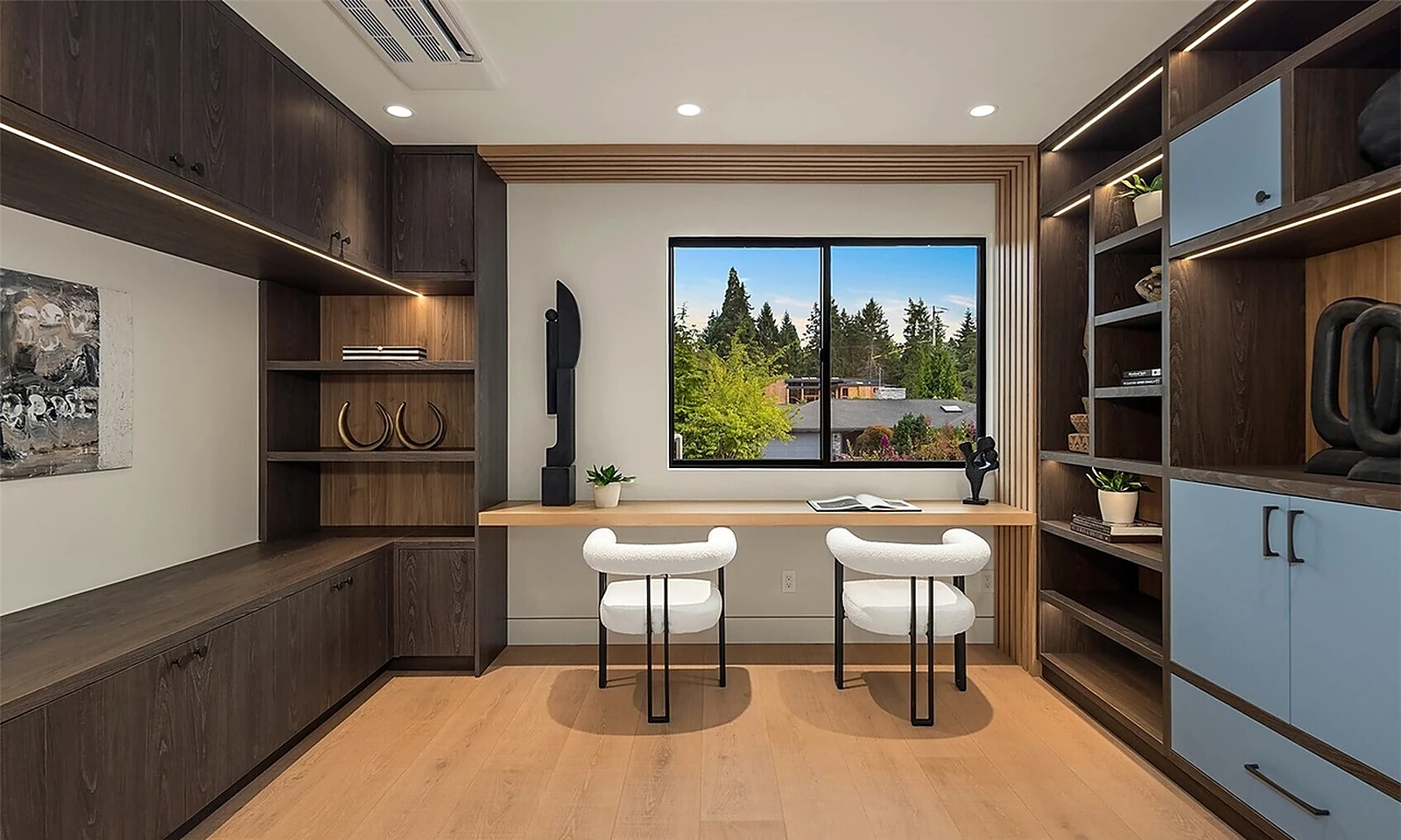 Study with dark wood built-ins, a floating oak desk, two white boucle chairs, and a window looking out to tall evergreens