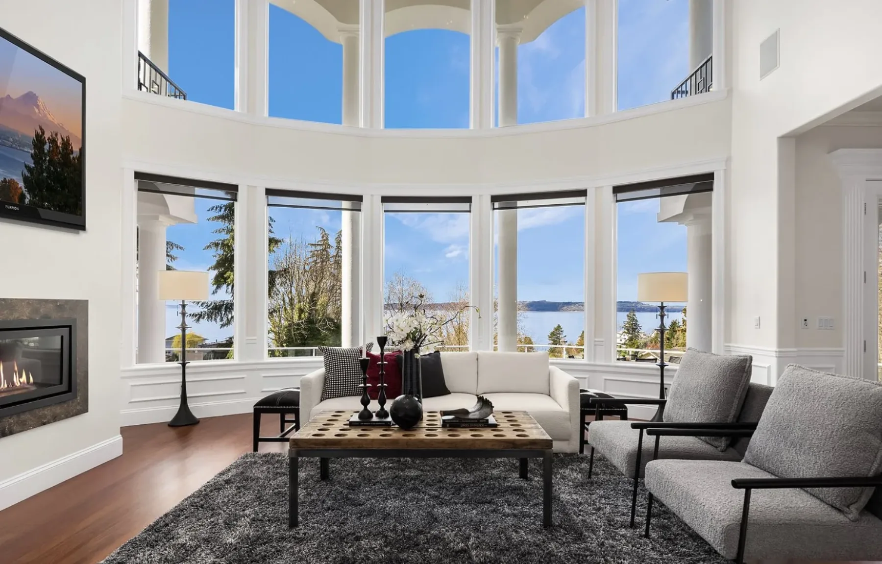 Two-story great room staged with curved walls of windows over Puget Sound, a linear fireplace, neutral seating, and a sculptural coffee table