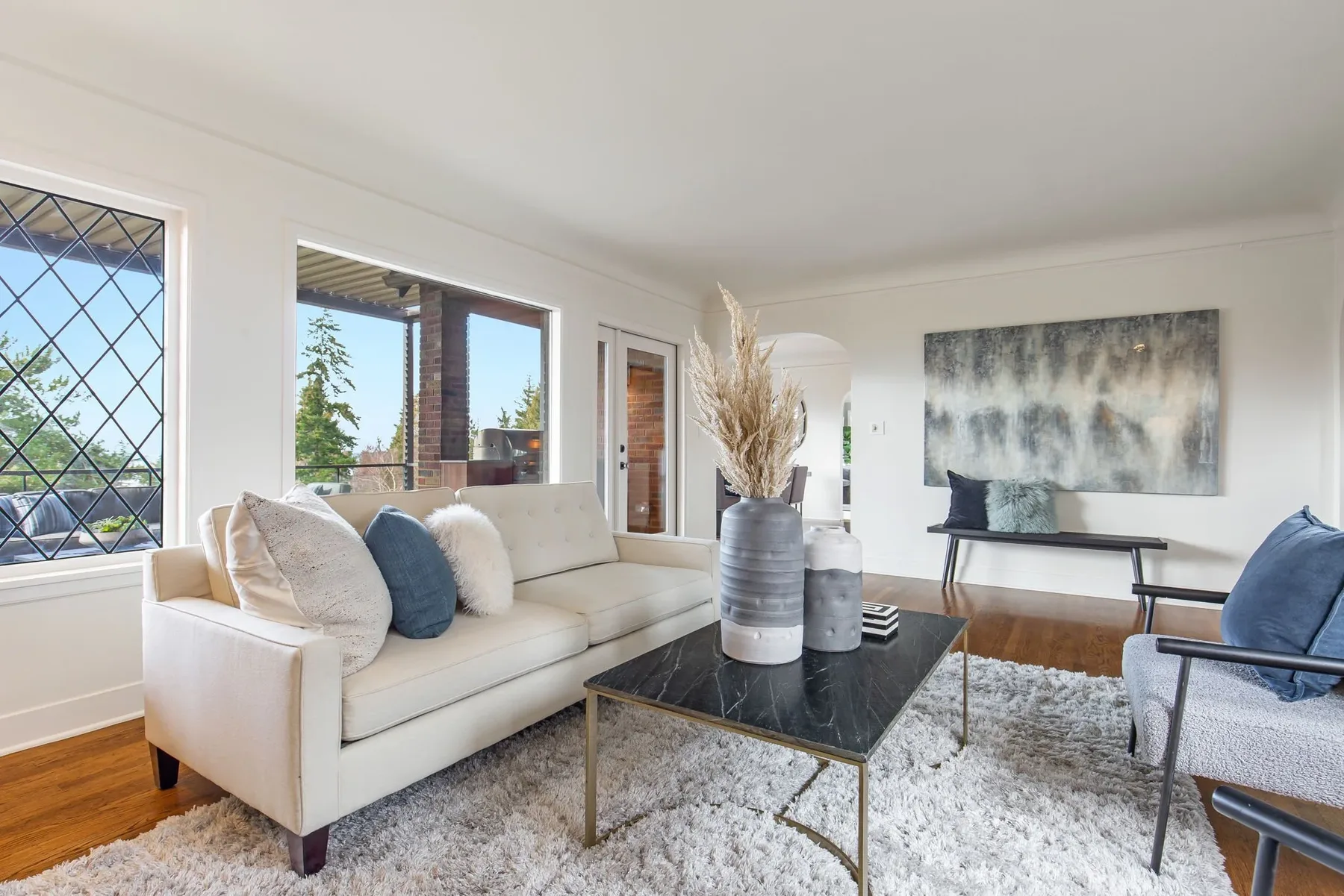 Living room sitting area staged with a white sofa, marble coffee table, abstract art, and a diamond-paned window framing the trees
