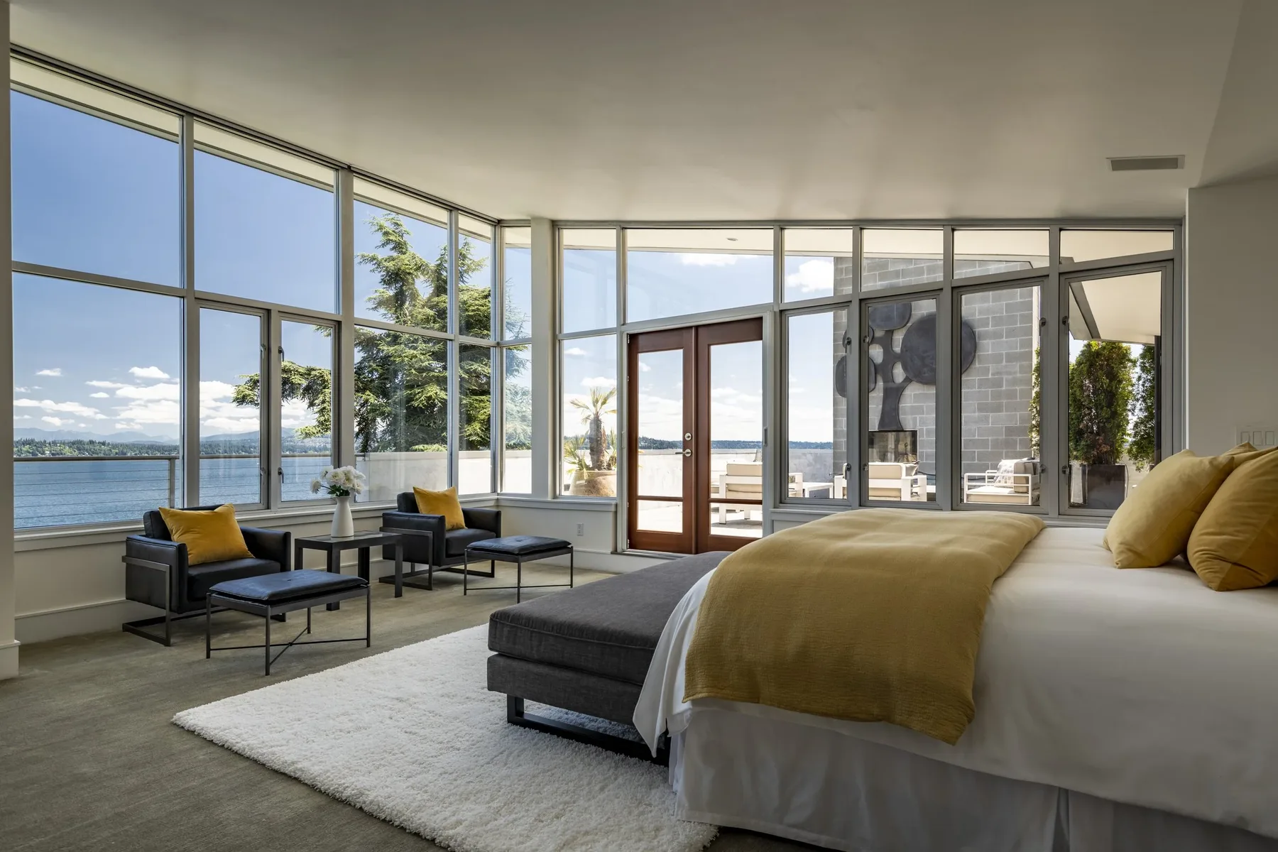 Primary suite staged with floor-to-ceiling corner windows, a gold throw, and steel-framed chairs framing sweeping Lake Washington views