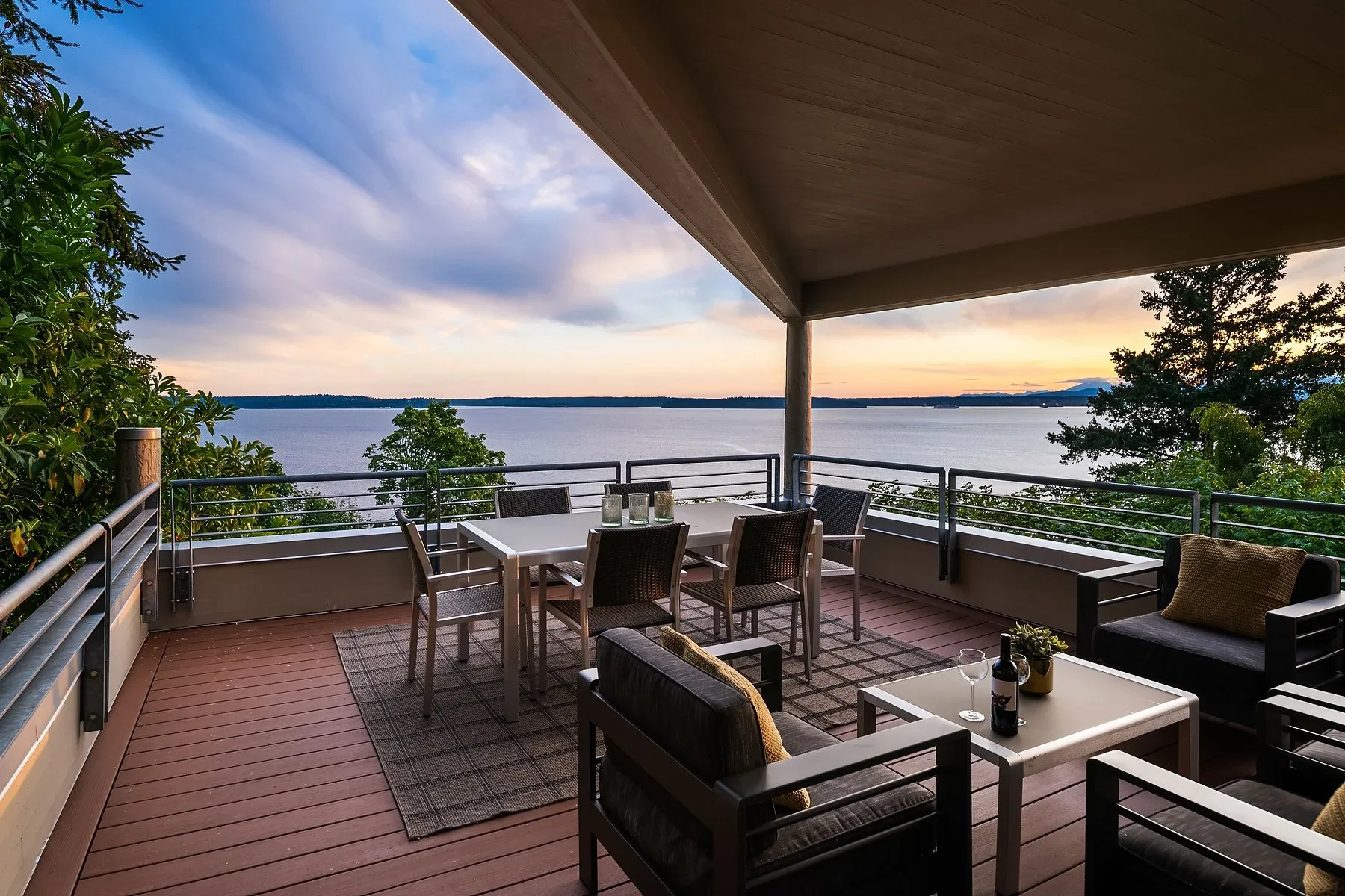 Covered waterfront deck staged with a dining set and lounge chairs overlooking Puget Sound at sunset