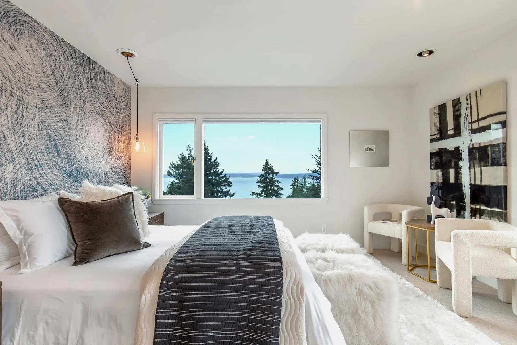 Primary bedroom staged with a swirl-pattern accent wall, crisp white bedding, and a window framing Puget Sound beyond the treetops