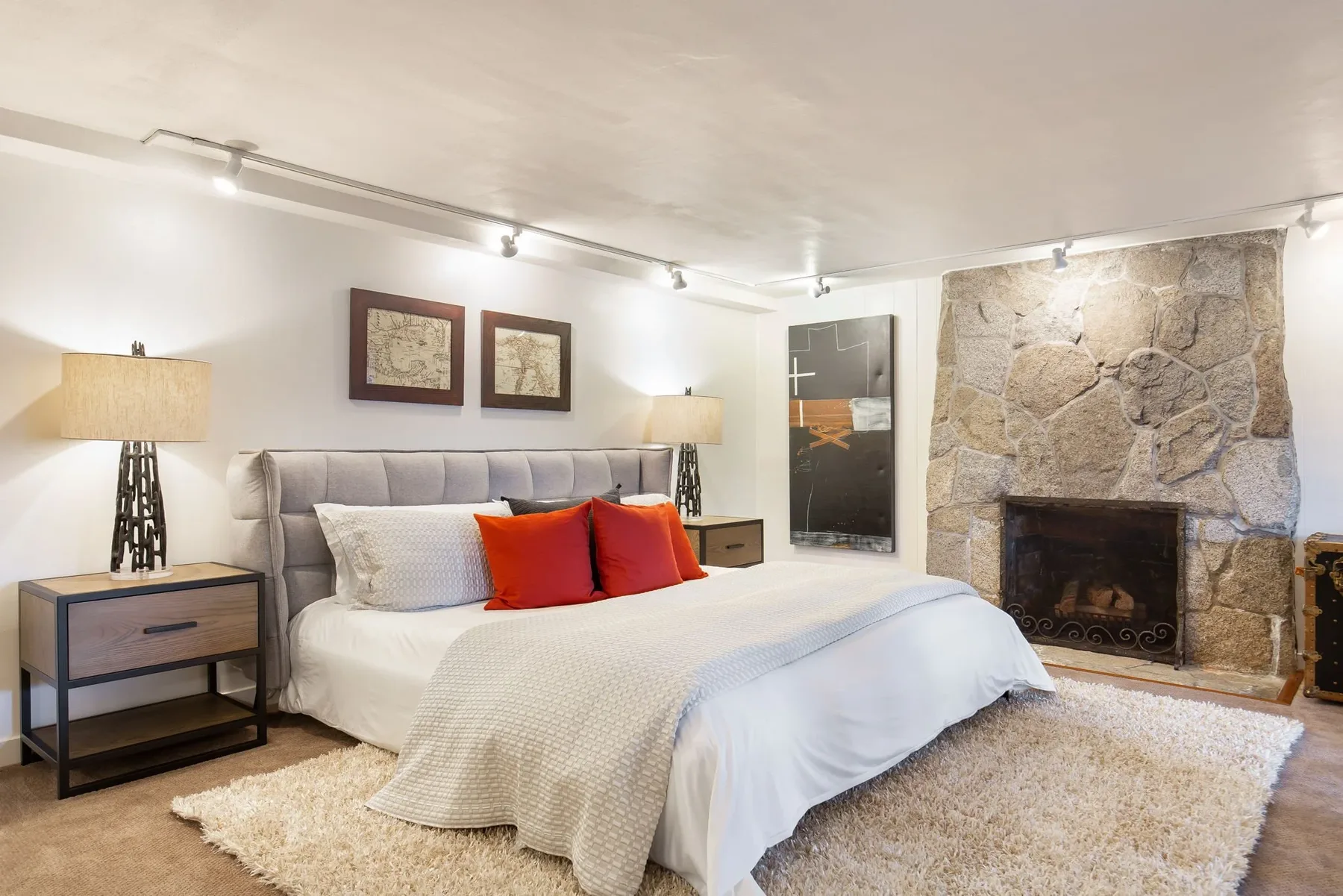 Bedroom staged with a gray tufted headboard, red accent pillows, and a floor-to-ceiling stone fireplace