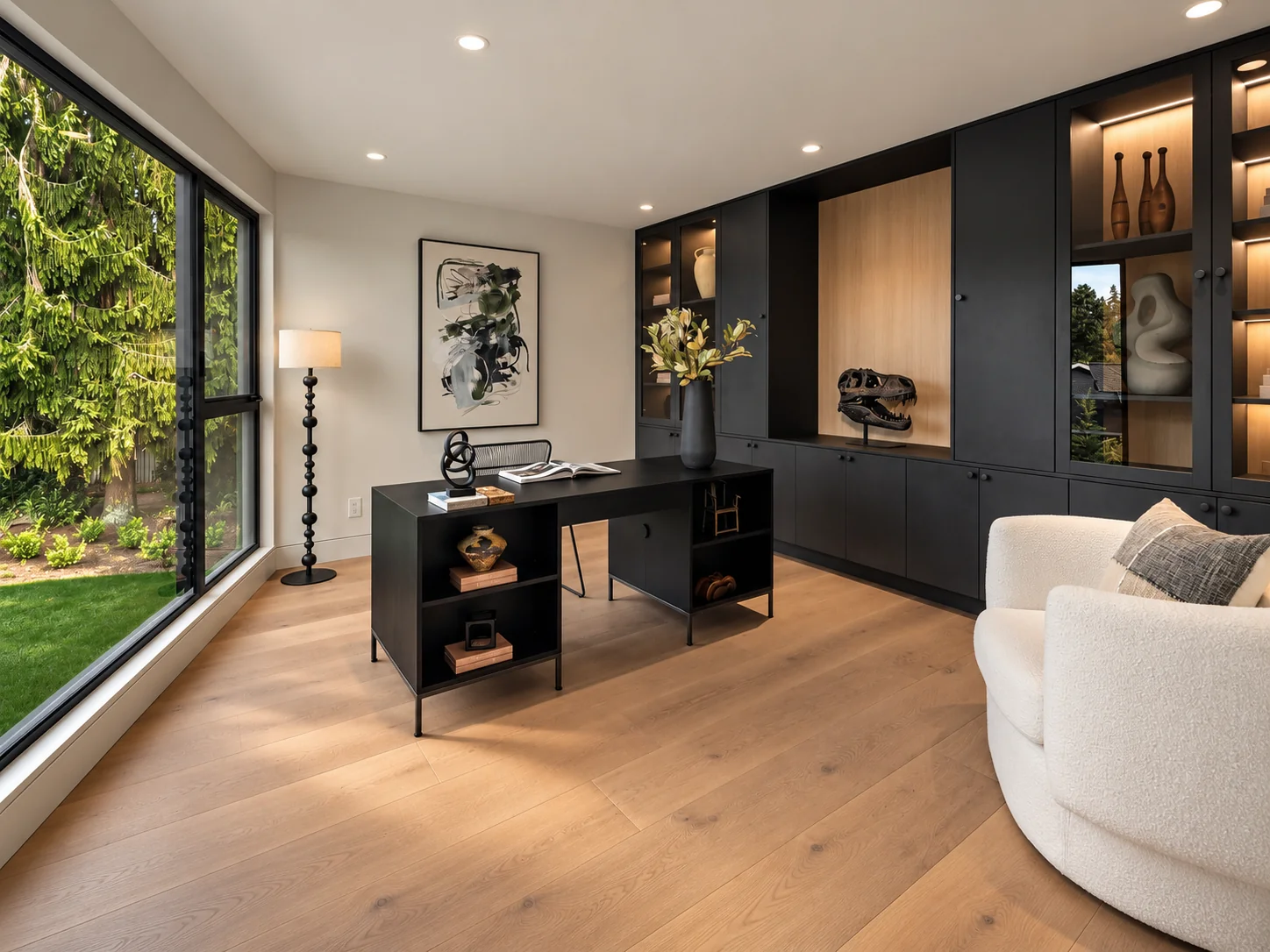 Luxury home staging in Clyde Hill featuring a modern home office with a black desk, lit built-in cabinetry, and a glass wall framing evergreen forest views.