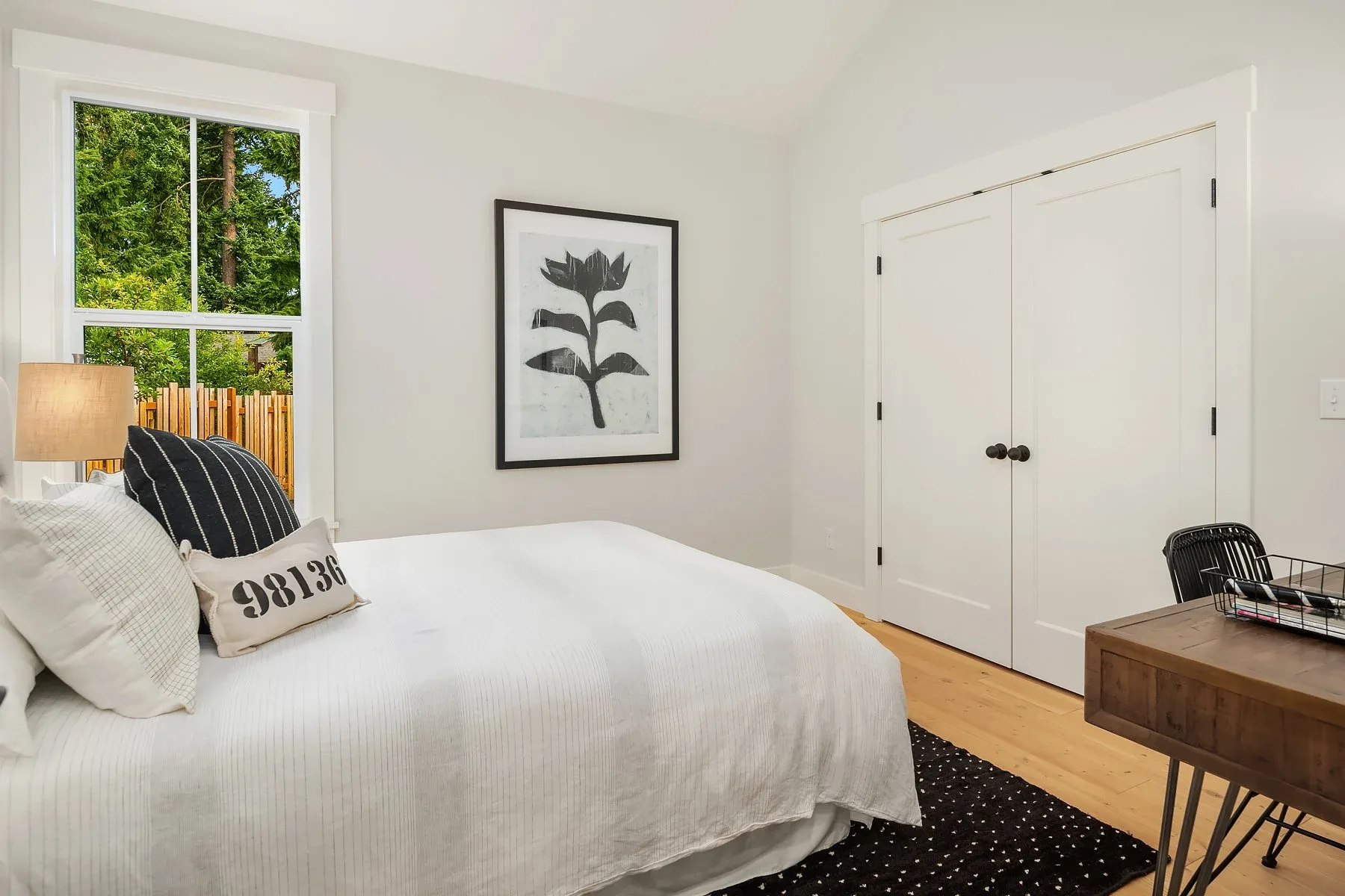 Guest bedroom staged with crisp white bedding, a botanical print, and a wood desk beside a window framing the trees