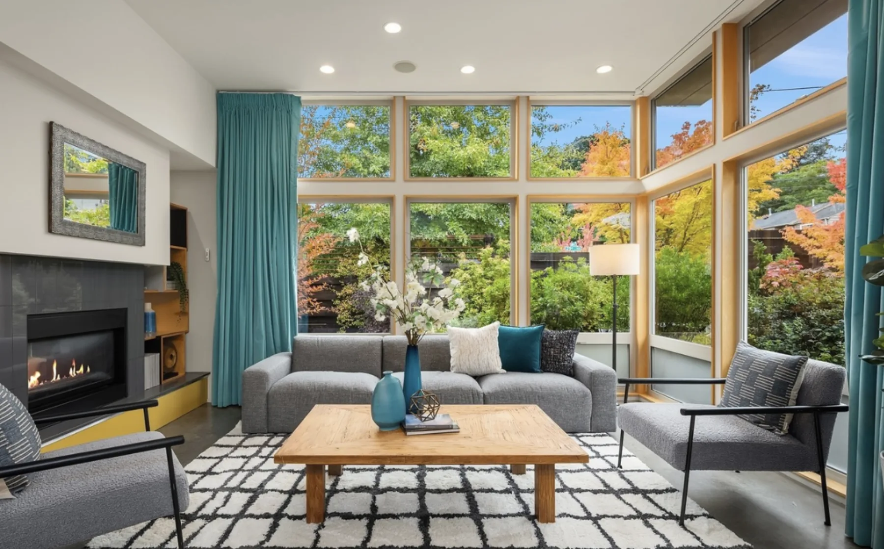 Living room staged by RD Interiors with a gray sectional, linear fireplace, teal curtains, and walls of wood-framed windows to the garden