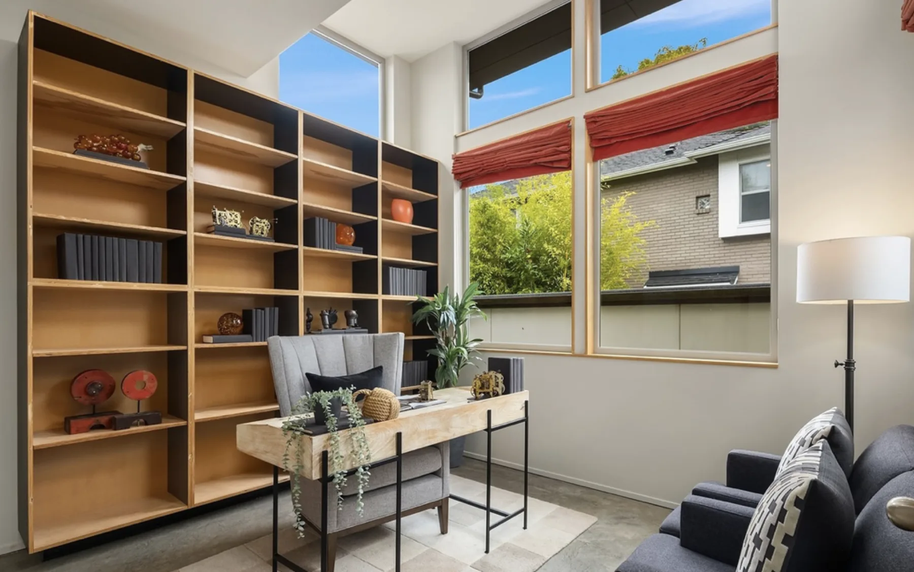 Study staged with floor-to-ceiling wood bookshelves, a live-edge desk, a gray chair, and tall windows with rust-colored shades