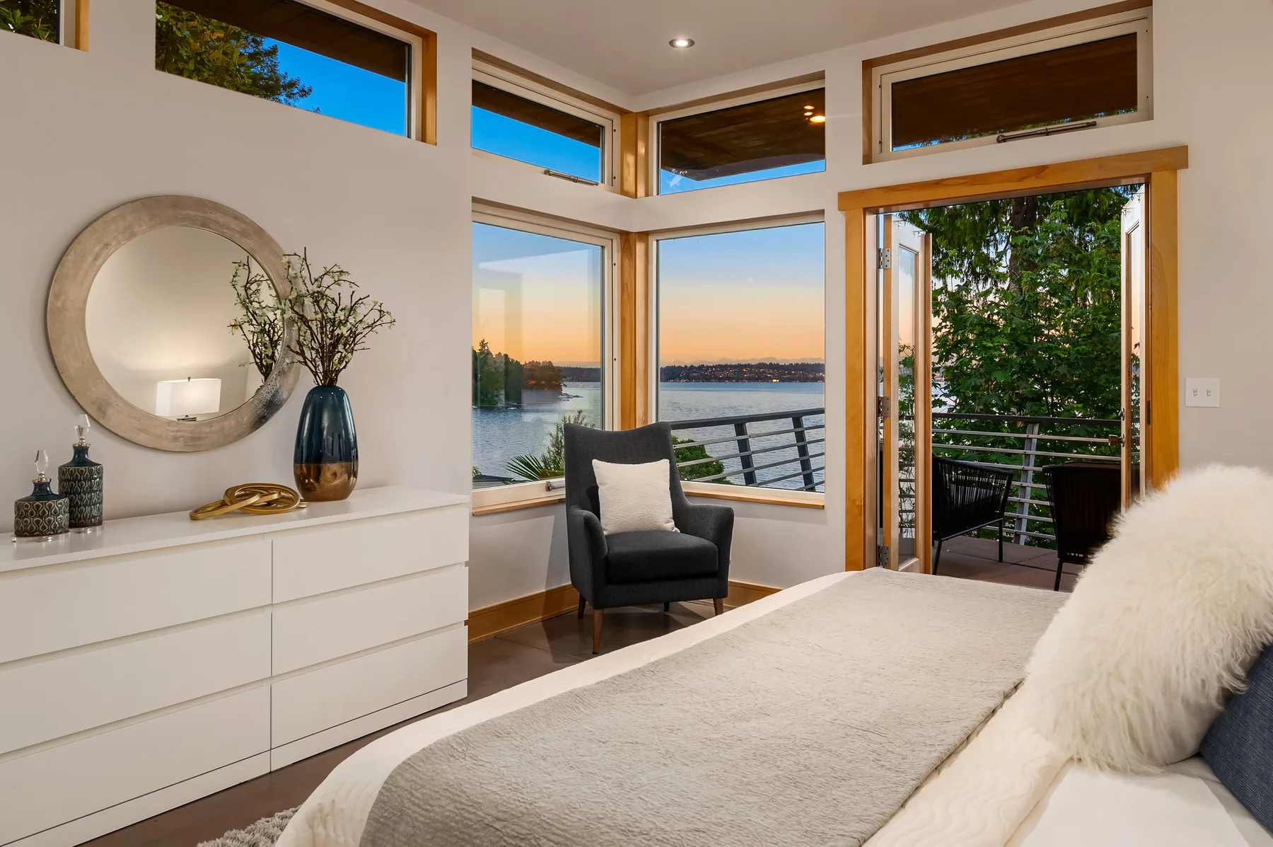 Primary suite staged by RD Interiors with a low dresser, round mirror, and corner windows framing the water at dusk