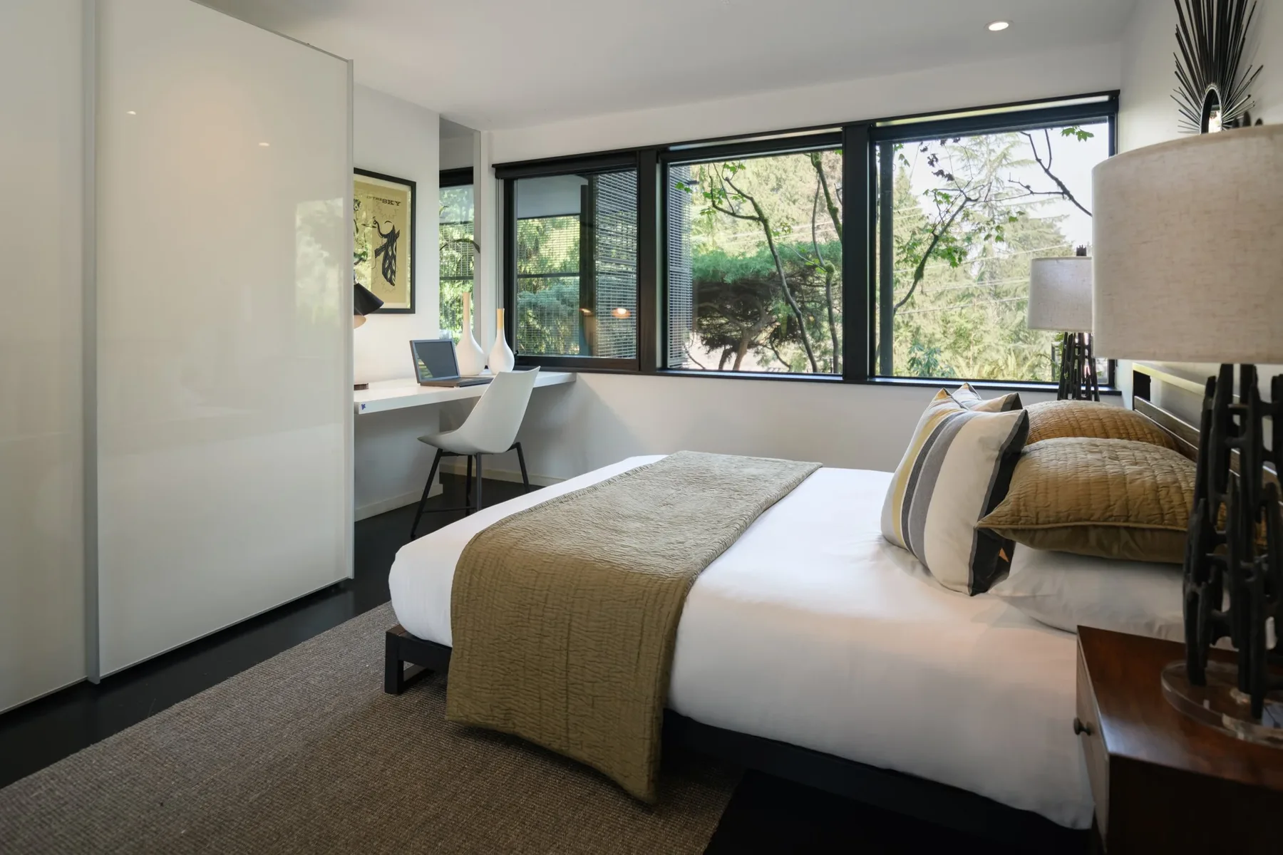 Modern primary bedroom staged by RD Interiors with a neutral upholstered bed, layered textiles, and treetop views