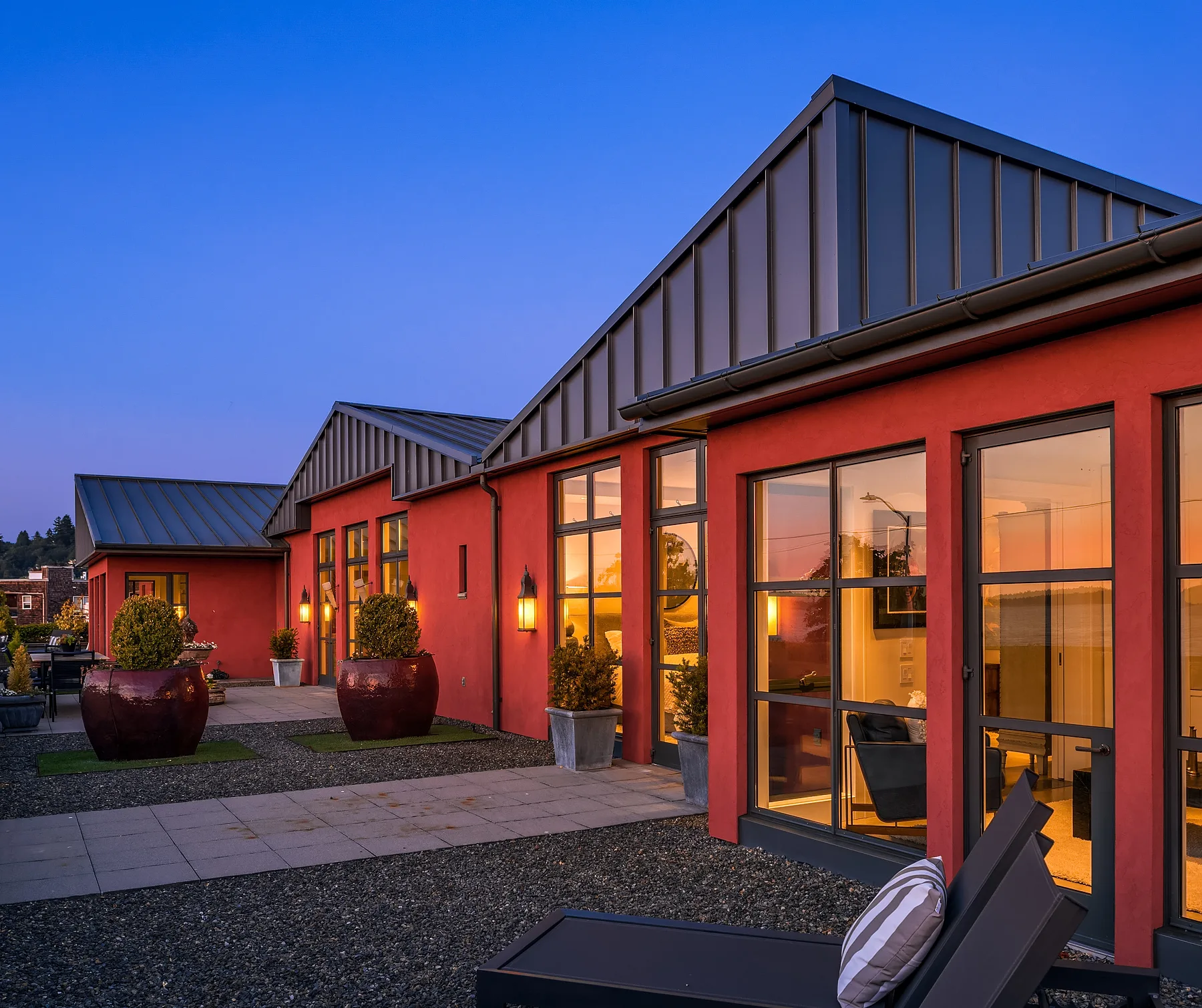 Terracotta-clad waterfront home at dusk with a standing-seam metal roof, glass doors, and a gravel courtyard