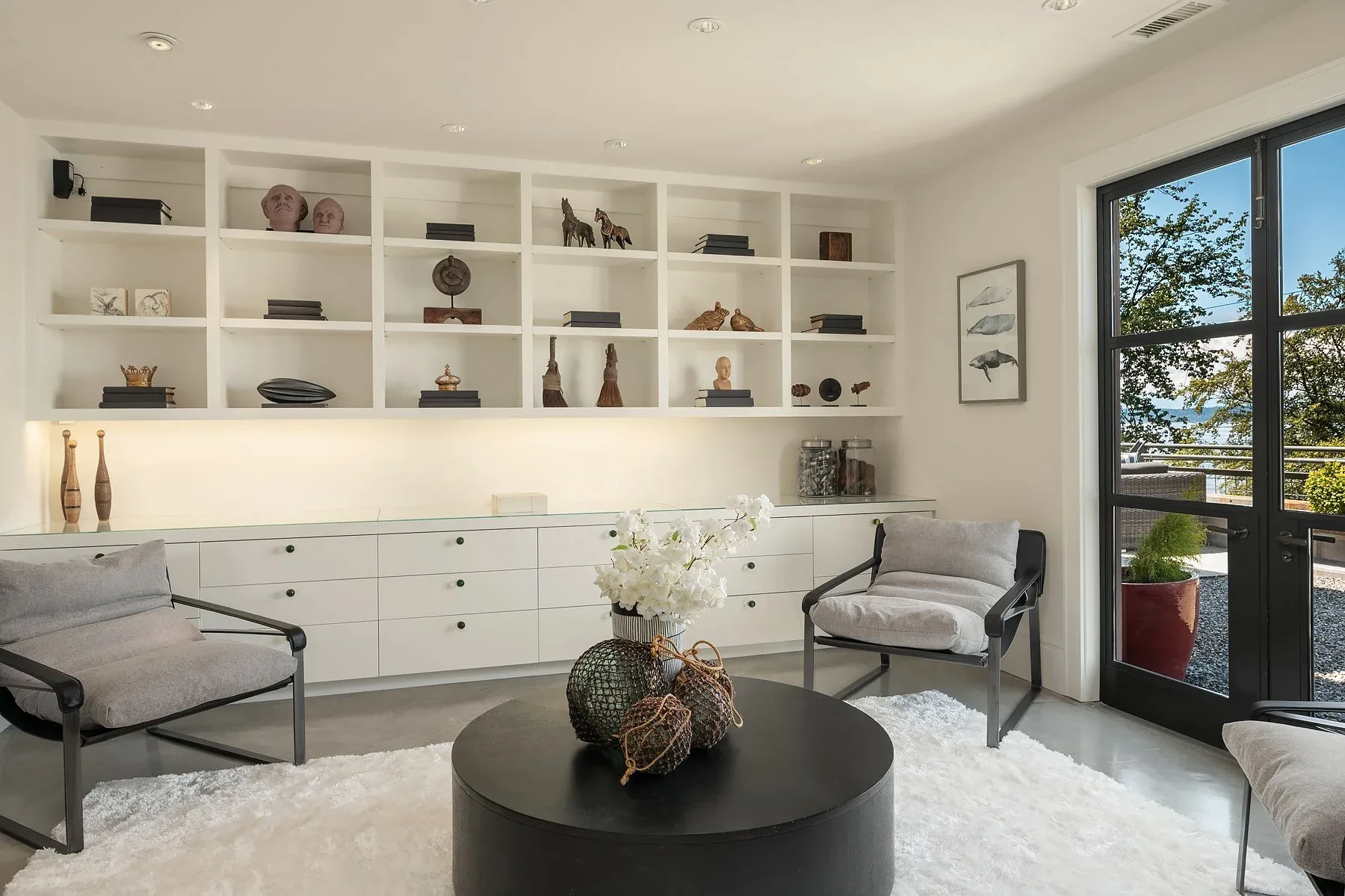 Study staged with white built-in shelving, gray sling chairs, a round black coffee table, and a door to the waterfront terrace