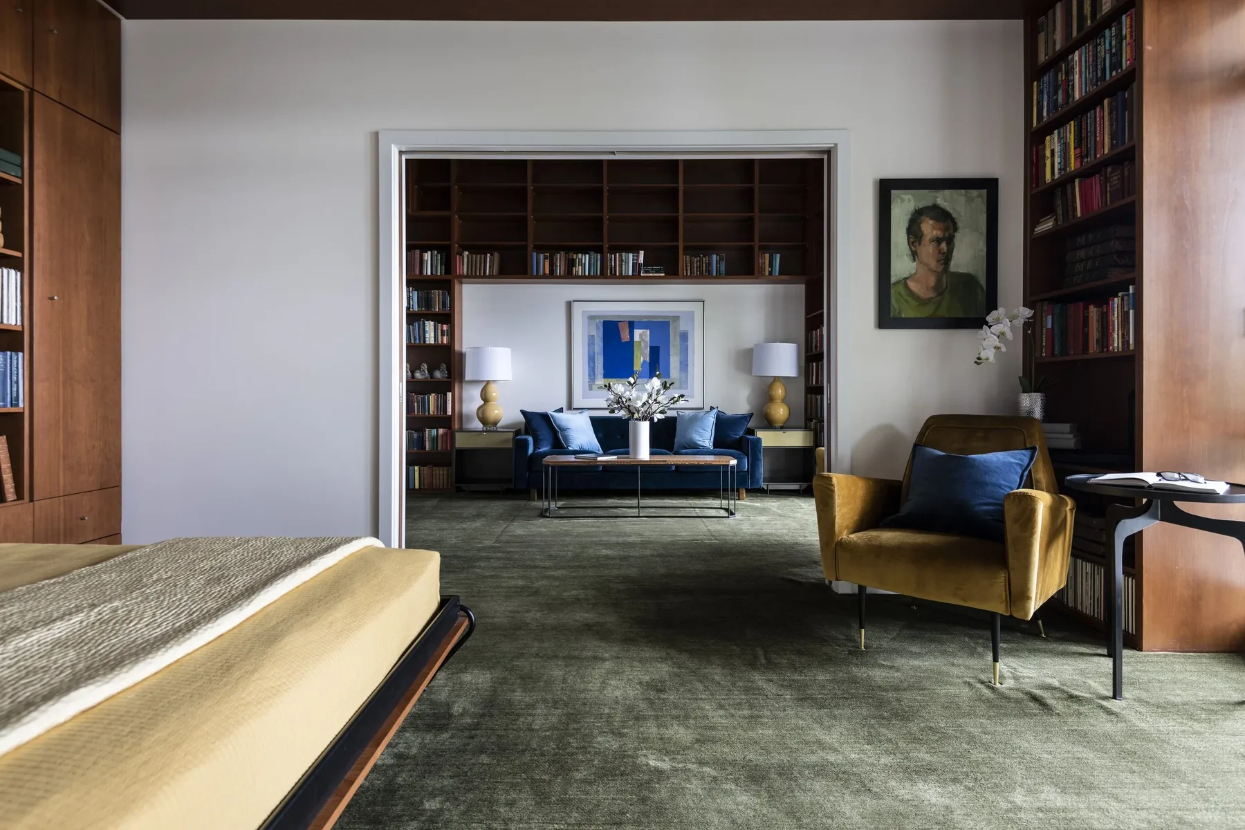 Bedroom staged with a green velvet carpet, gold accent chair, and a wood-paneled library beyond with a blue settee and built-in shelving