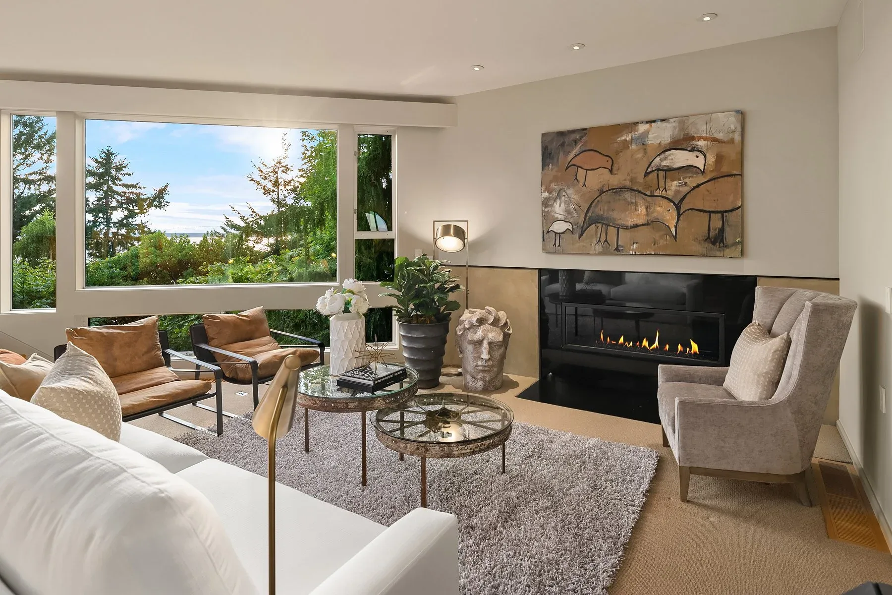 Living room staged with a linear fireplace, bird artwork, tan leather chairs, and a picture window framing the trees and water