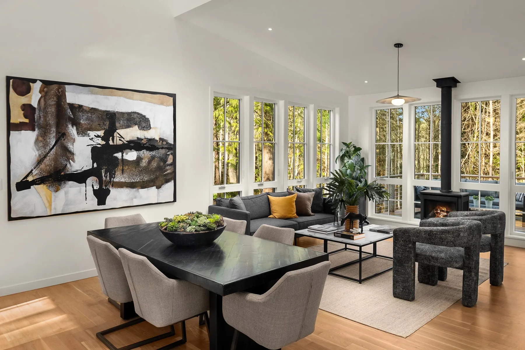Open great room staged by RD Interiors with a dining table, neutral seating, a wood stove, and walls of windows to the forest