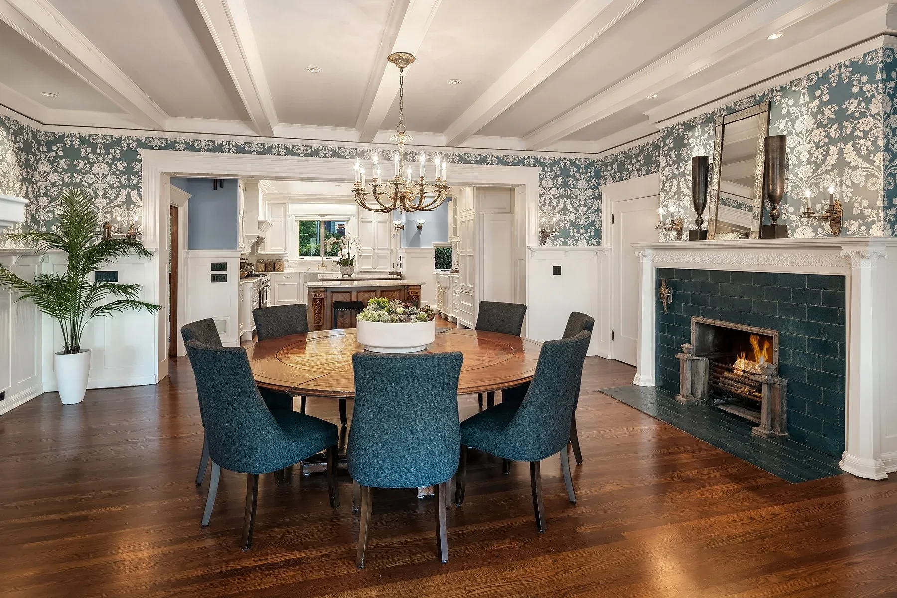 Dining room staged by RD Interiors with a round wood table, blue upholstered chairs, patterned wallpaper, and a blue-tiled fireplace beside a coffered ceiling