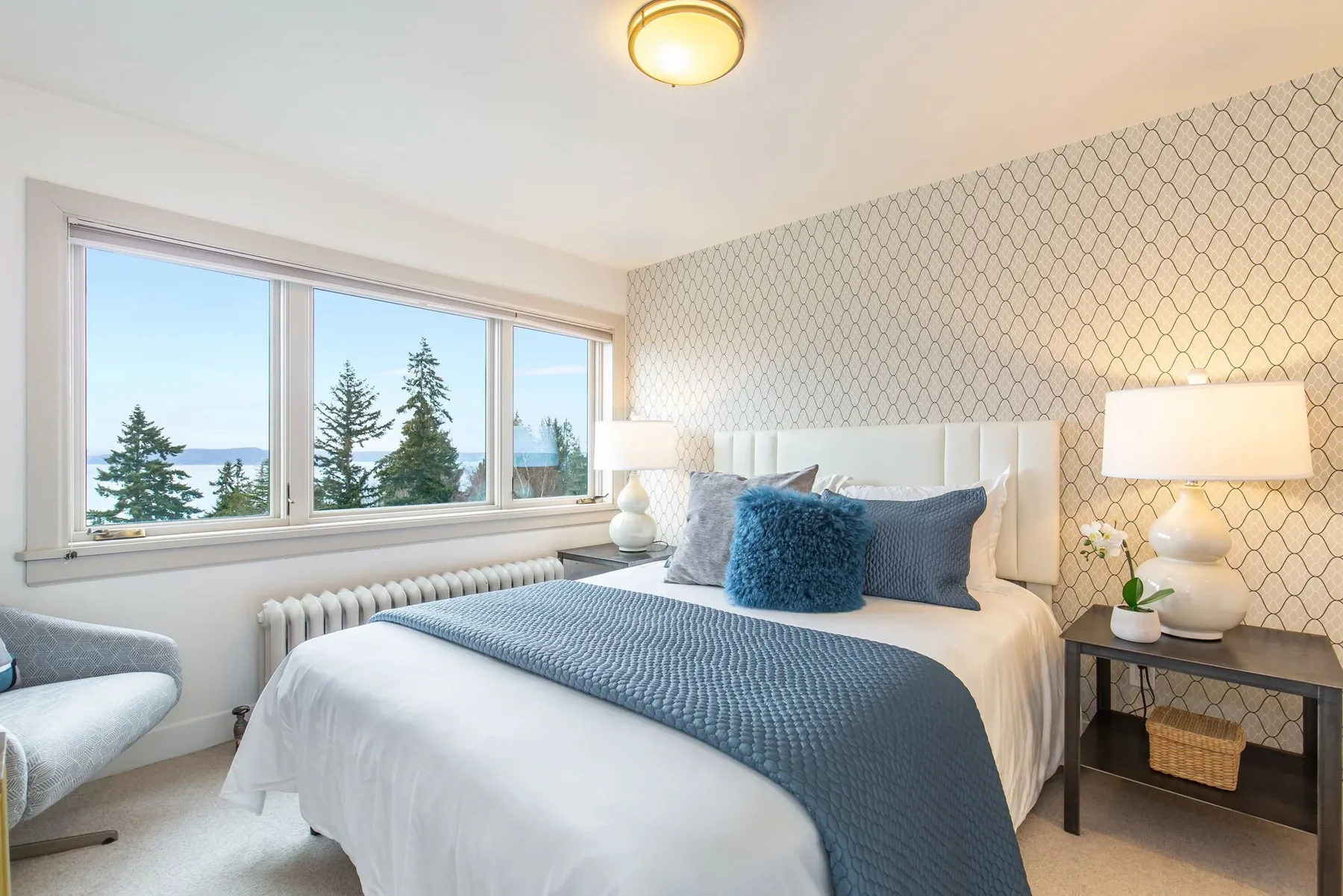 Bedroom staged by RD Interiors with a white upholstered headboard, blue layered bedding, trellis wallpaper, and a Sound view through the window