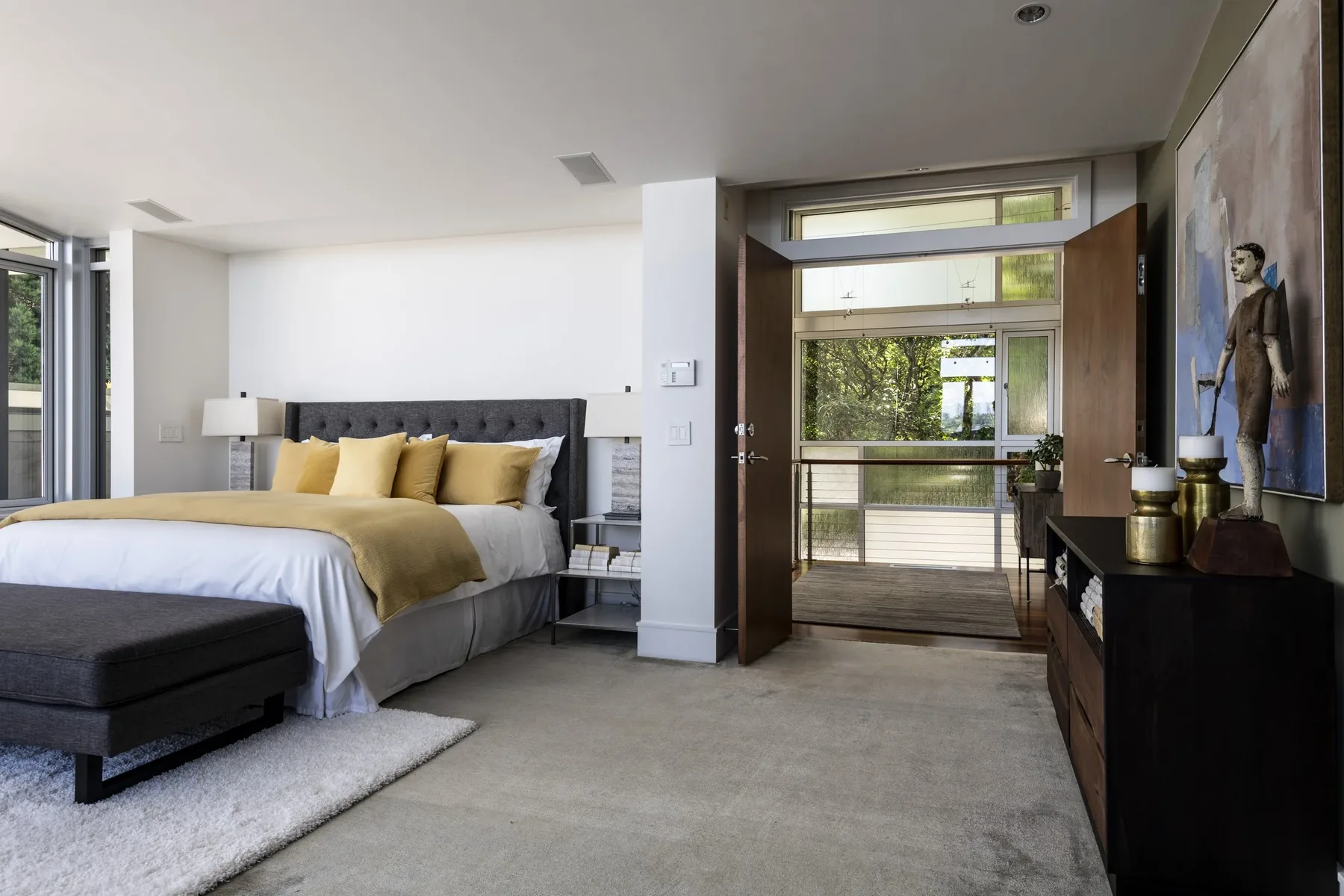 Primary bedroom staged by RD Interiors with a tufted gray headboard, gold accent pillows, and a door opening to a terrace among the trees