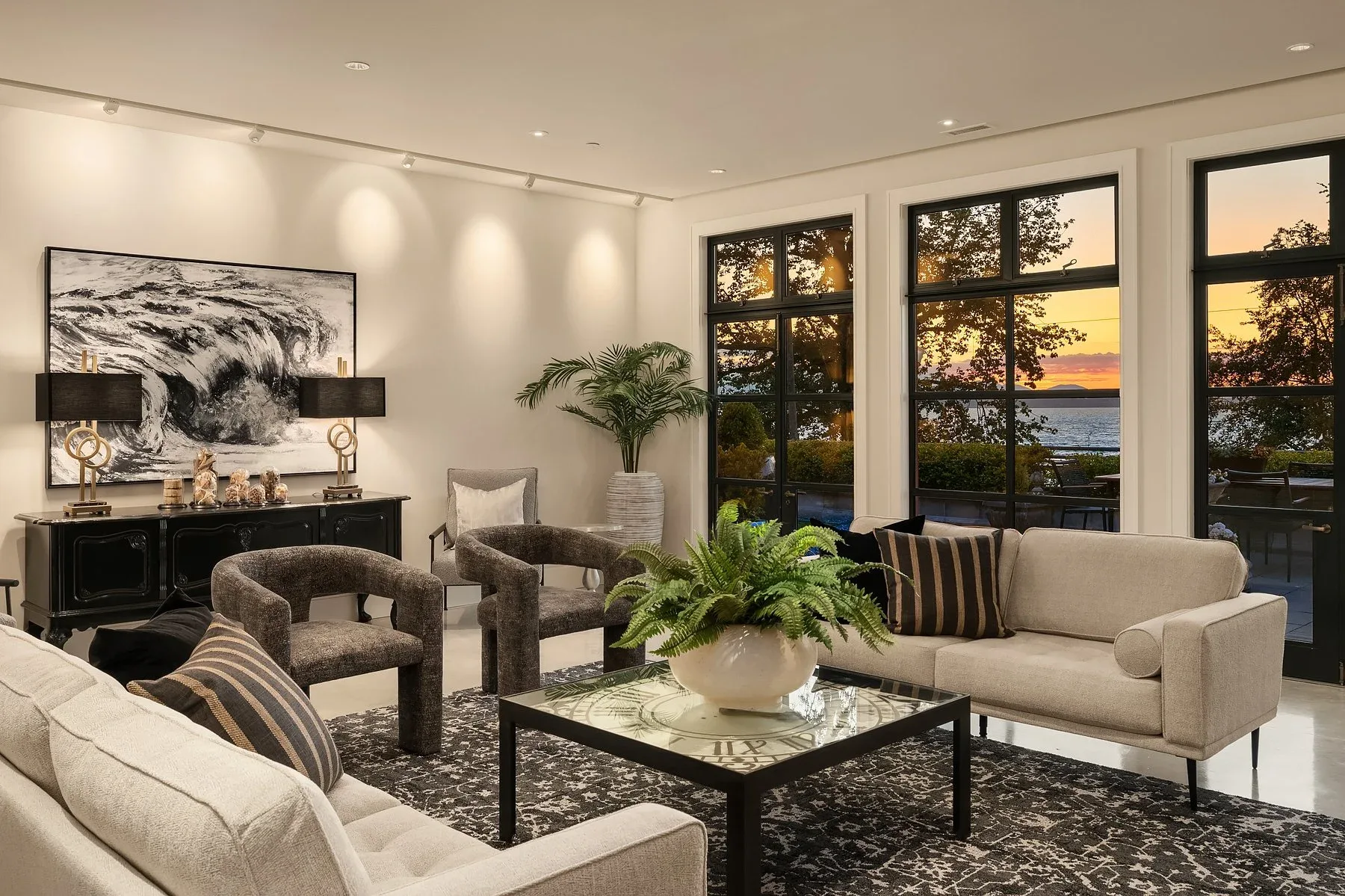 Seattle luxury home staging featuring a fireside sitting area at dusk with sculptural chairs, a wave painting, and water views through black-framed windows