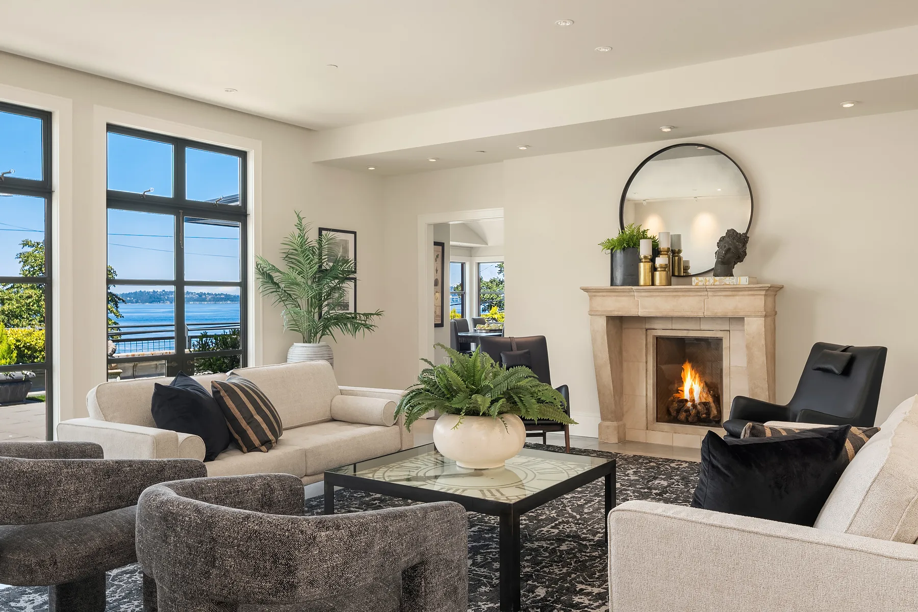 Living room with a limestone fireplace, round mirror, neutral sofas, and water views through floor-to-ceiling windows