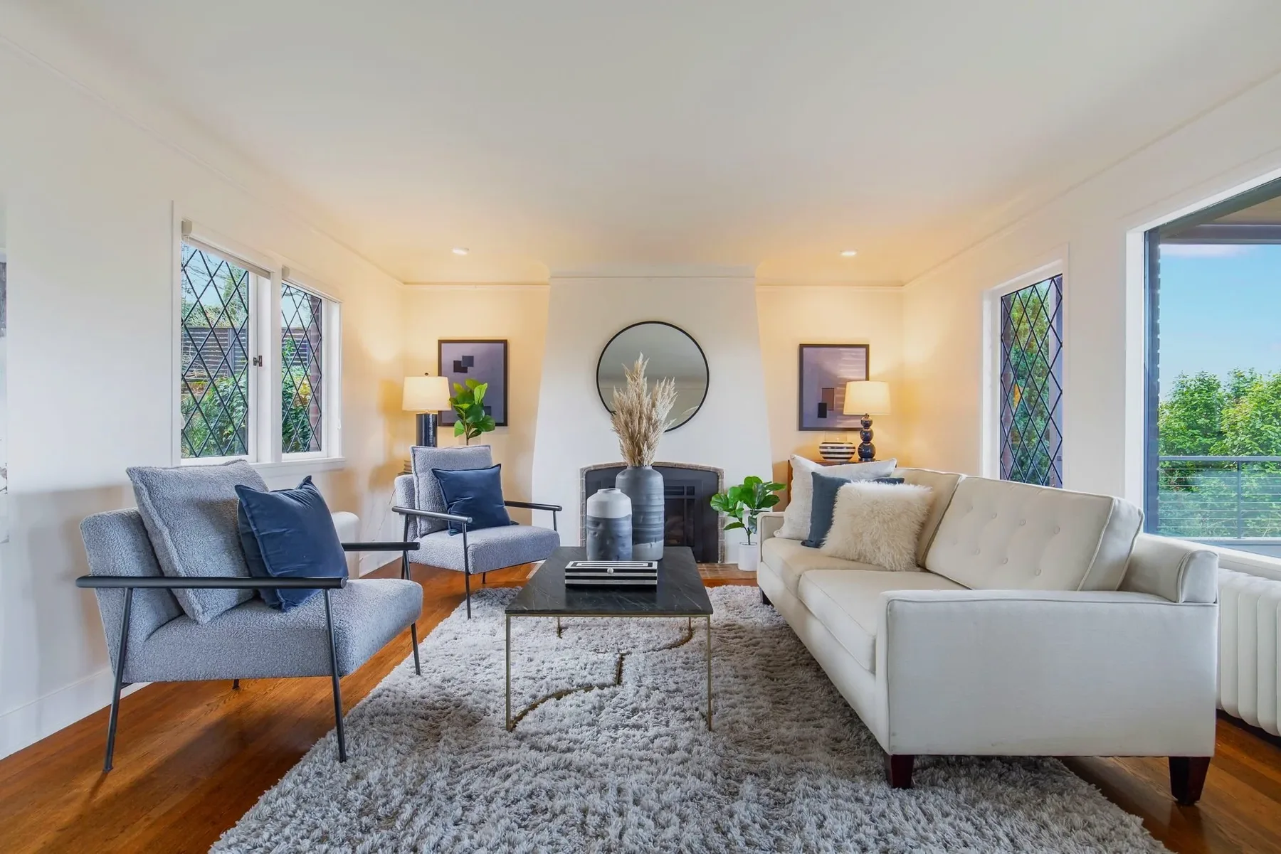 Living room staged by RD Interiors with a white tufted sofa, blue accent chairs, a round mirror over the fireplace, and leaded glass windows