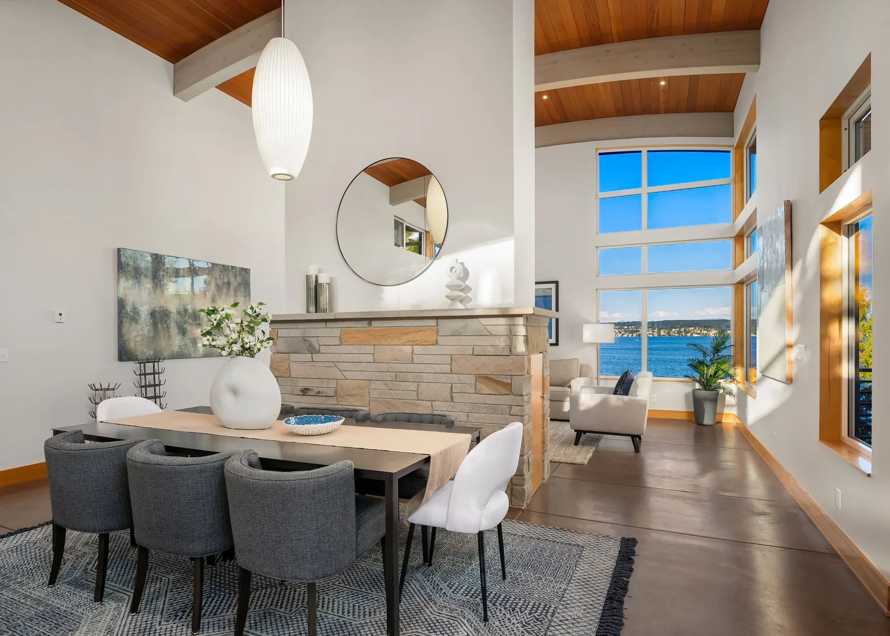 Contemporary dining room staged with upholstered chairs beside a stacked-stone fireplace and walls of glass to the lake