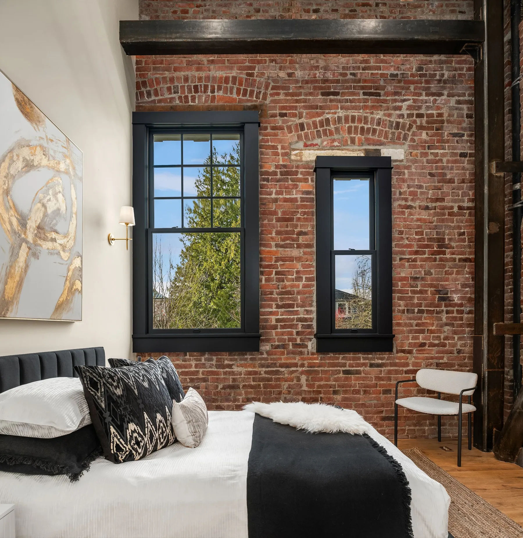 Bedroom staged with layered neutral bedding against an exposed brick wall and tall steel-framed windows