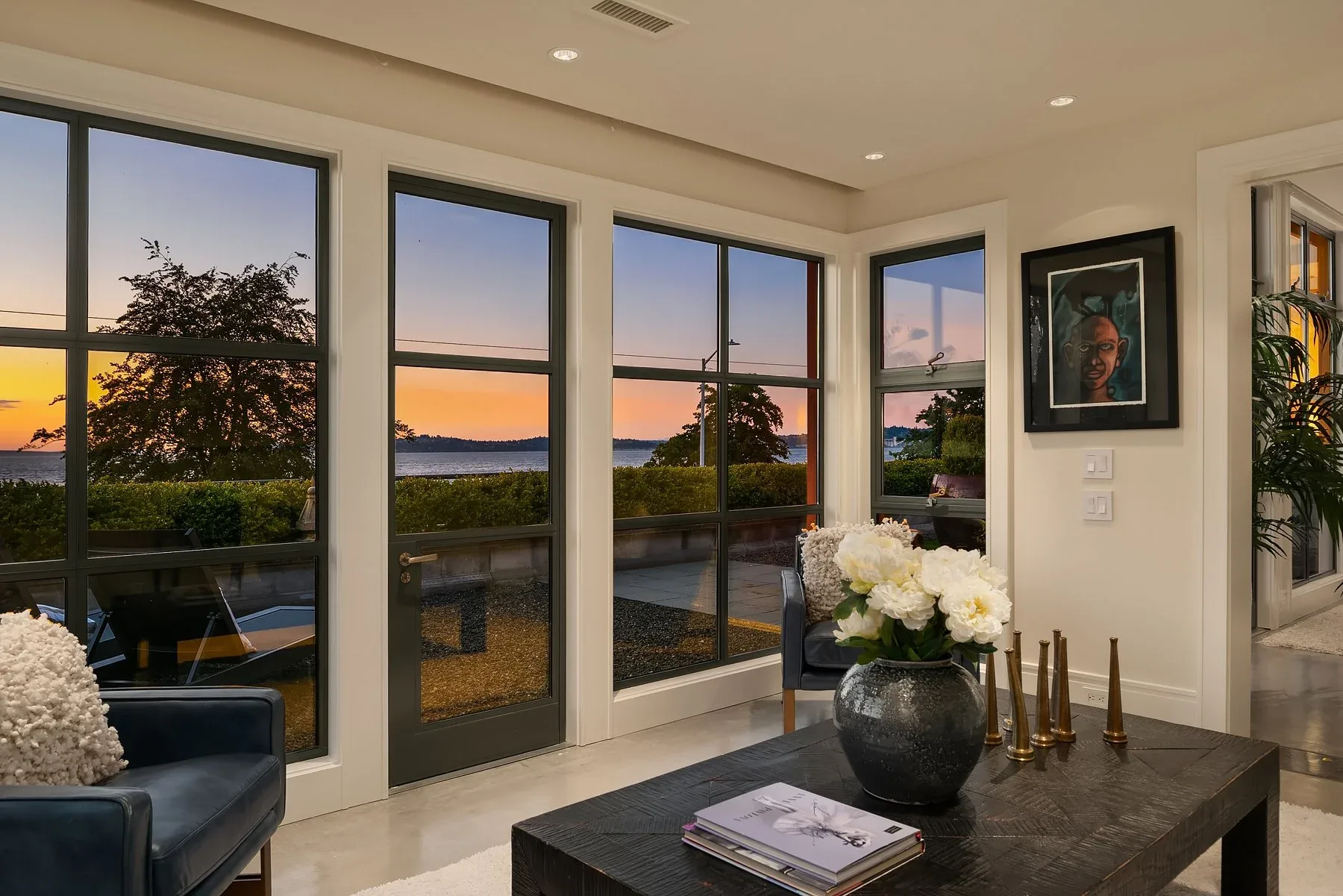 Corner sitting room staged with a console, blue leather chairs, fresh peonies, and walls of windows over a Puget Sound sunset