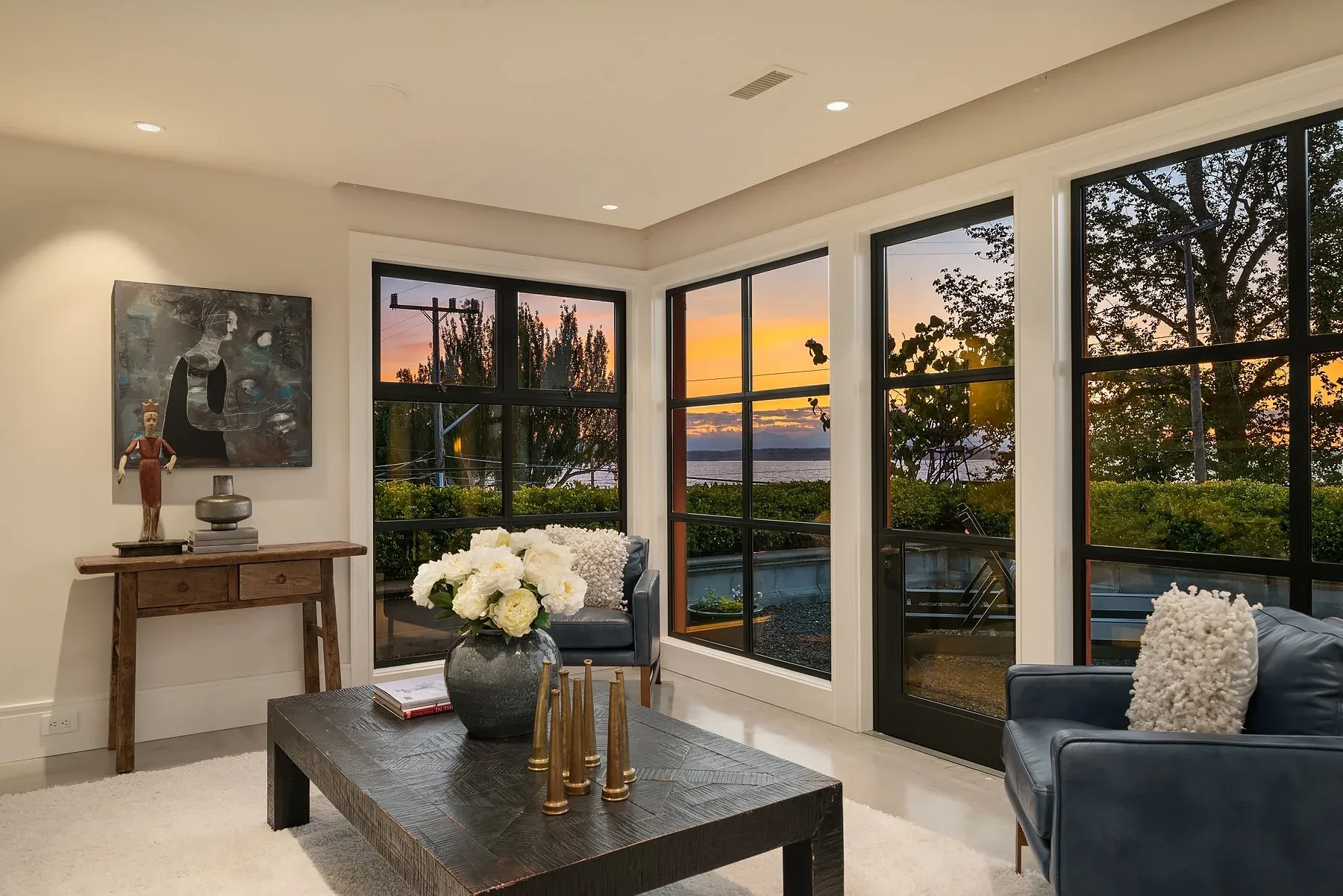 Sitting nook staged by RD Interiors with a wood coffee table, blue leather chairs, and steel-framed windows framing the sunset water view