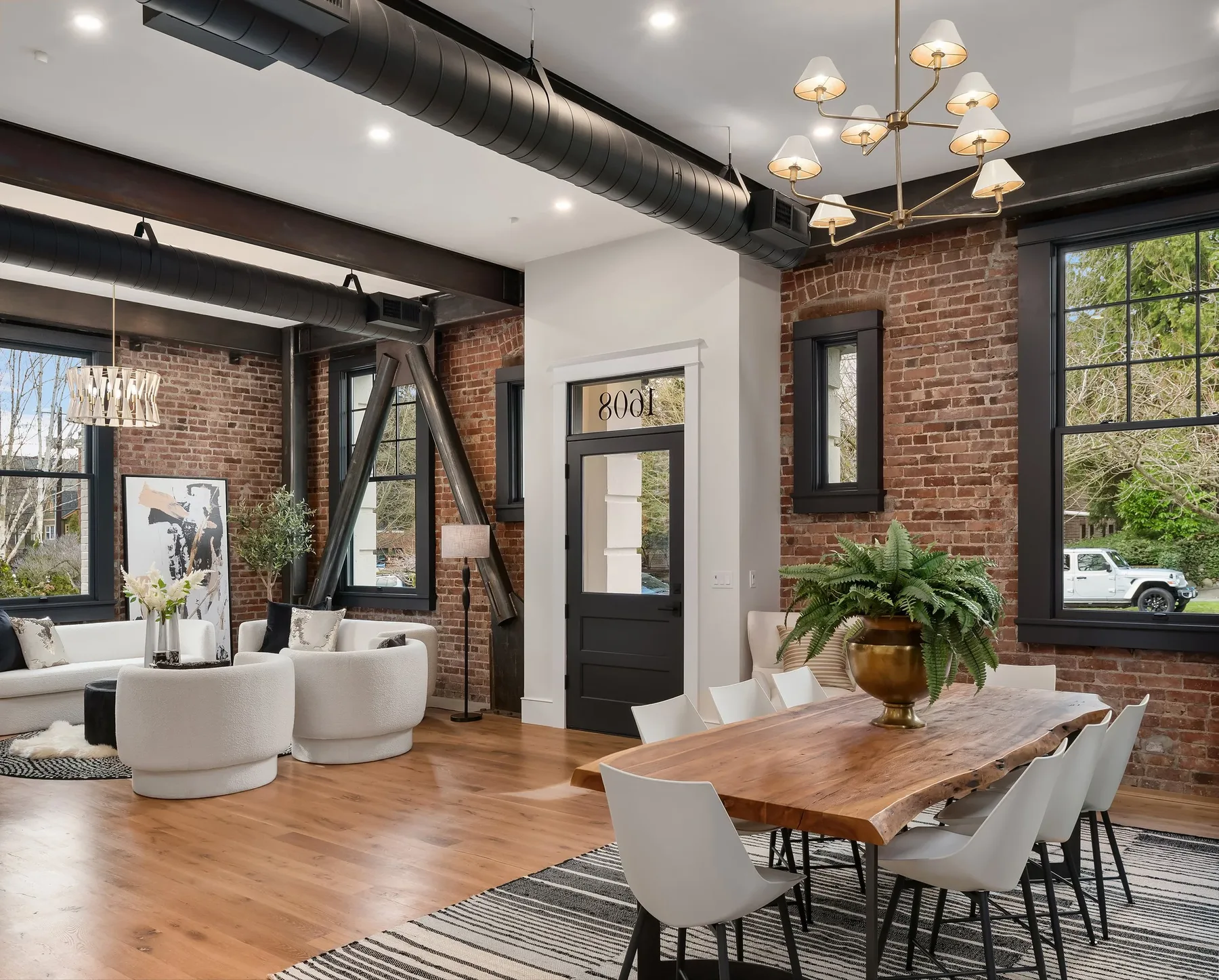 Open living and dining area beneath original brick and steel, staged with bouclé seating and a live-edge table