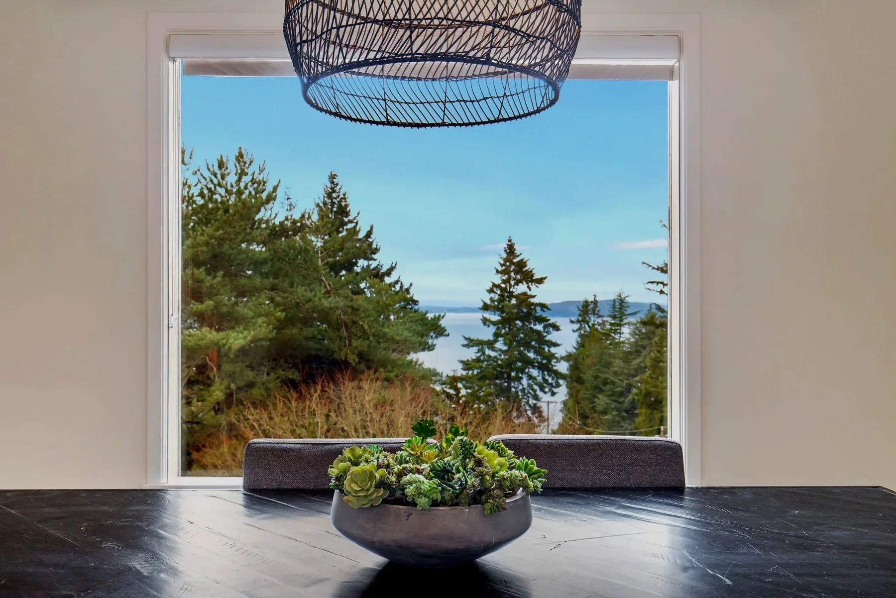 Dining table styled with a succulent centerpiece and woven pendant beneath a picture window framing evergreens and Puget Sound