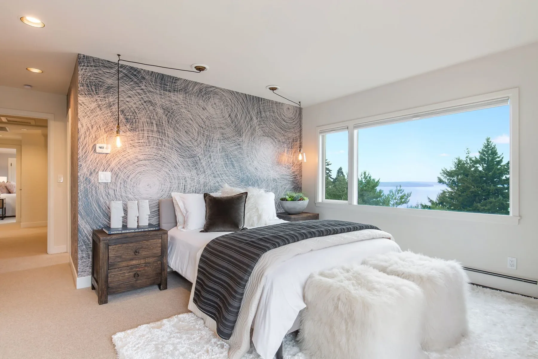 Primary suite staged with a dramatic swirl mural wall, wood nightstands, a sheepskin bench, and corner windows over the water