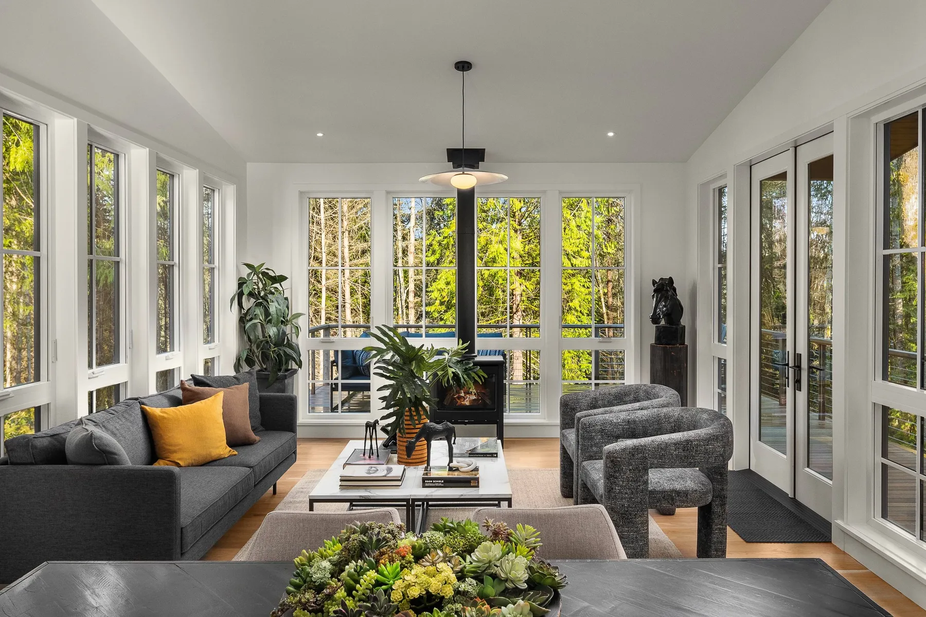 Living room staged with a vaulted ceiling, walls of windows, a wood stove, and bouclé accent chairs overlooking the trees
