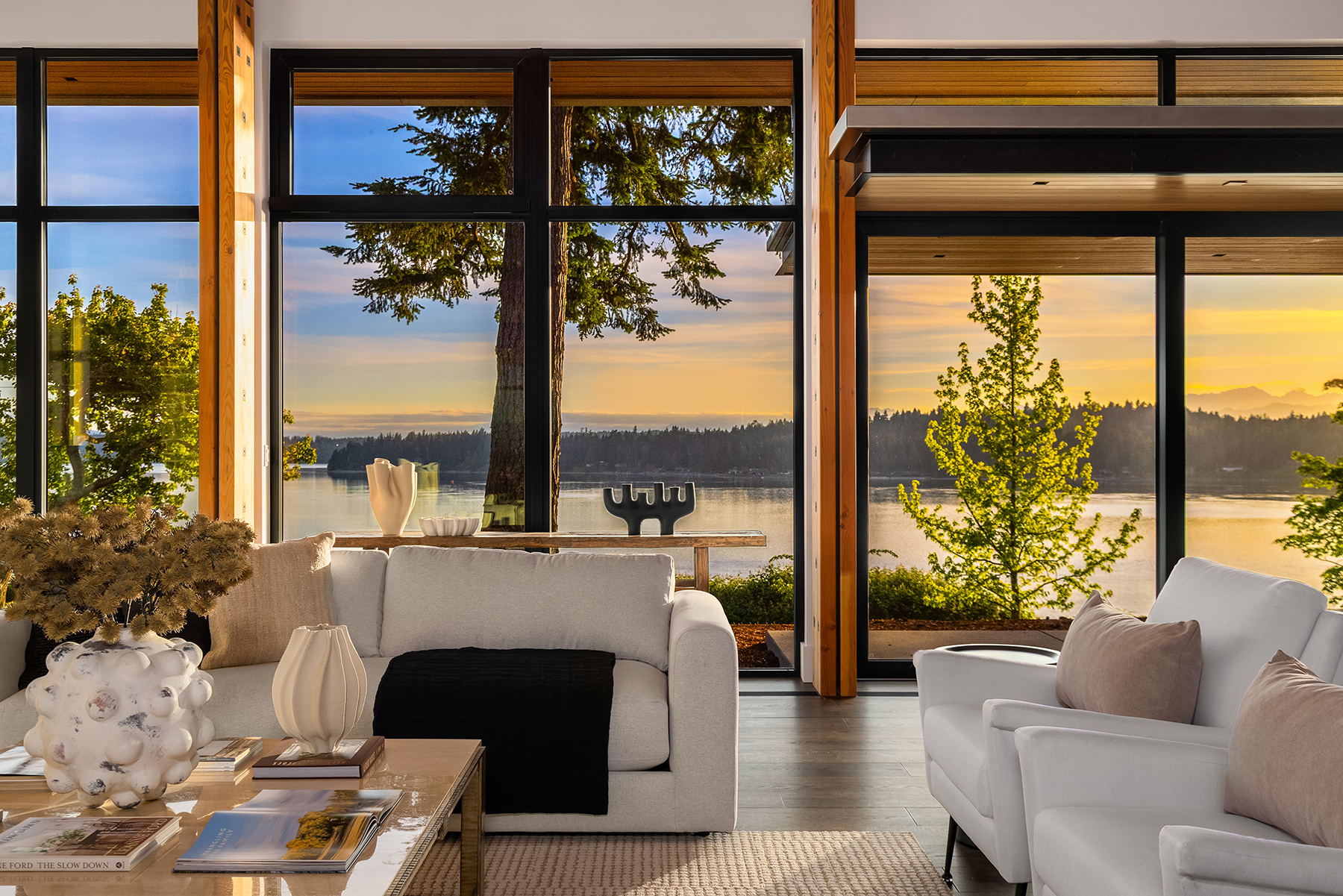Staged sitting room with soft white seating, sculptural decor, and wide windows overlooking the shoreline at dusk