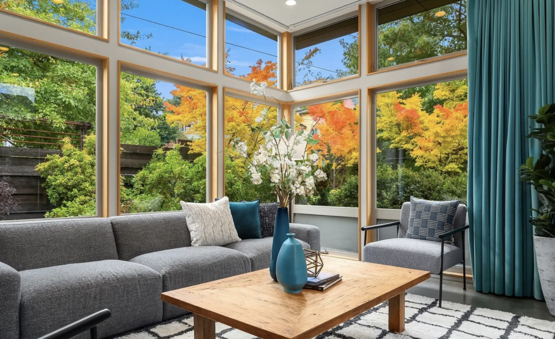 Seattle luxury home staging featuring a corner sitting area with a gray sectional, blue accents, and walls of windows framing fall foliage