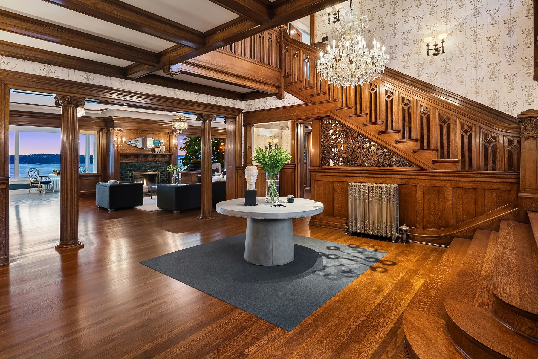Grand entry hall staged by RD Interiors with a carved wood staircase, crystal chandelier, fluted columns, and a round center table over water views