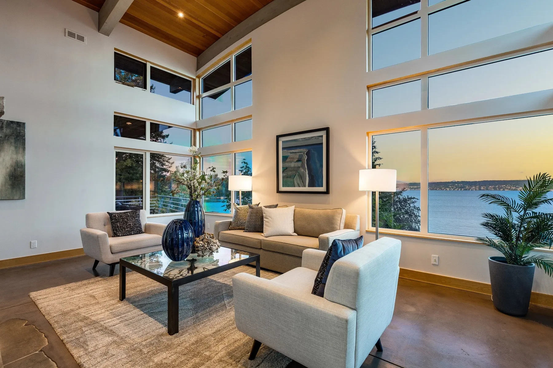 Lakefront living room staged by RD Interiors with floor-to-ceiling windows, a glass coffee table, and serene blue accents at sunset
