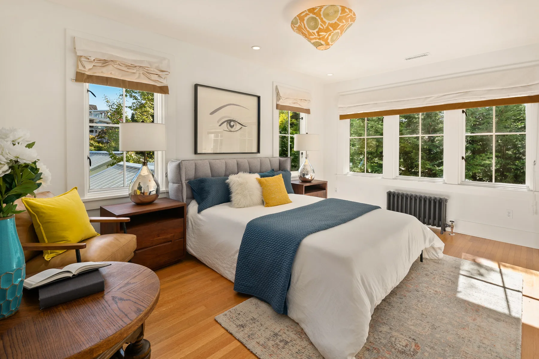 Primary bedroom staged with an upholstered gray bed, navy throw, gold accent pillows, and walls of windows framing the trees