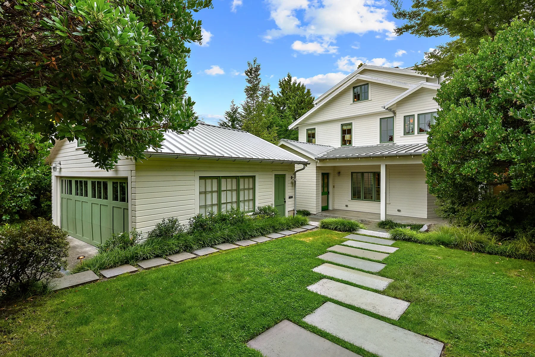 White craftsman lakehouse staged by RD Interiors with a green garage door, standing-seam metal roof, and a bluestone path across the lawn