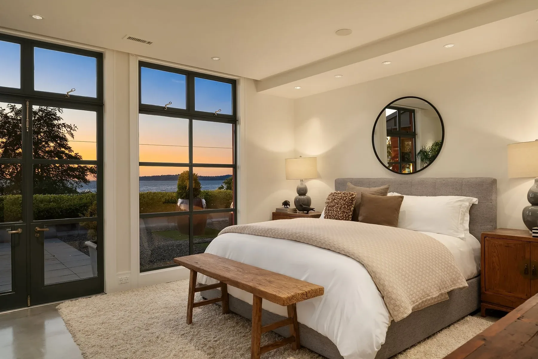Primary bedroom staged with a gray upholstered bed, round mirror, antique nightstands, and French doors opening to a Puget Sound sunset