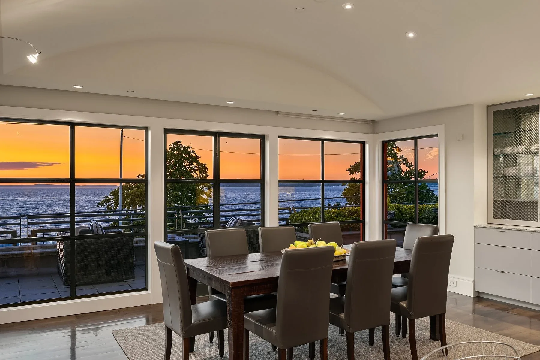 Dining room staged by RD Interiors with a wood table, leather chairs, and walls of steel-framed windows glowing over Puget Sound at sunset